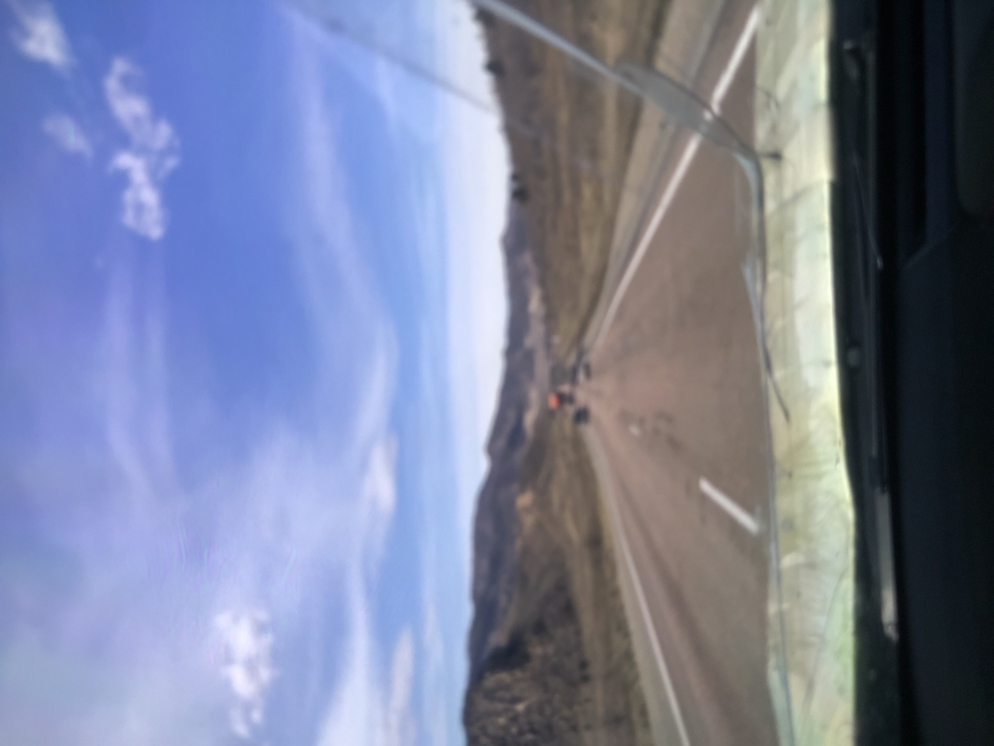 A blurred, vertically-oriented image showing a highway flanked by a mountainside under a partly cloudy sky, as seen from inside a vehicle, possibly a car. The road appears to be empty except for some indistinct dark shapes that are potentially other vehicles or people in the distance. The mountainside is arid, with dry vegetation. The vehicle's interior, likely part of the window or dashboard, is visible in the foreground.