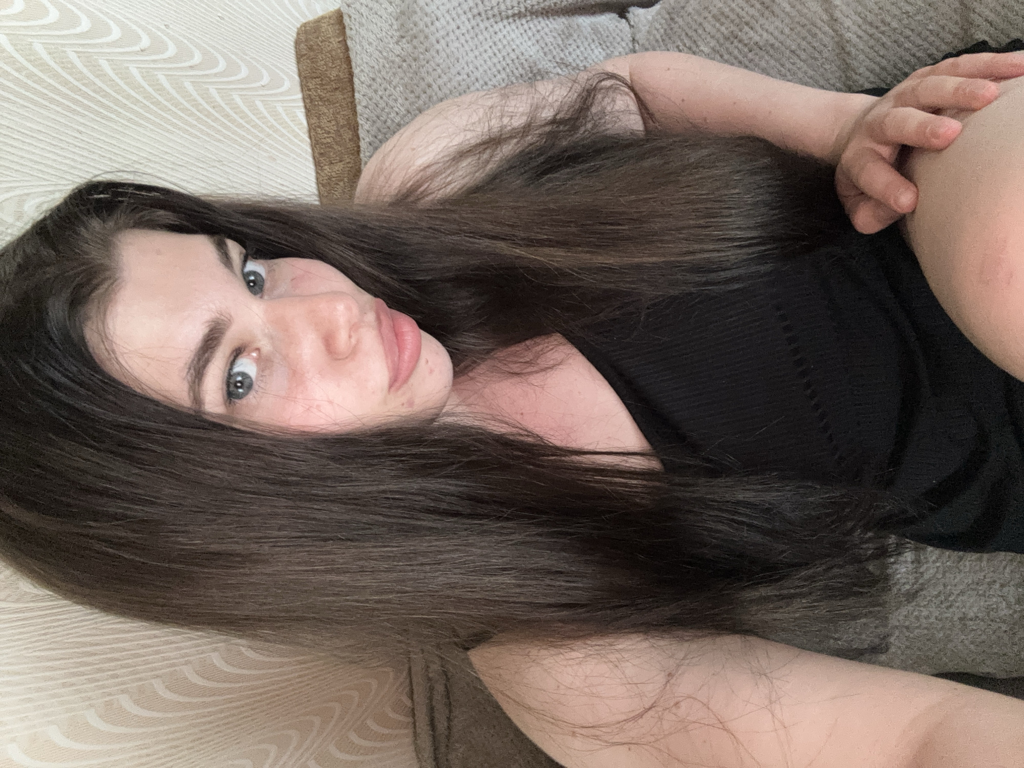 A woman with long, dark brown hair is lying on a beige patterned sofa or couch. She is wearing a black tank top. Her right hand is slightly raised near her right shoulder. Her face is visible, showing light skin, gray eyes, and pink lips. The background consists of the sofa and potentially a wall with a light-colored patterned wallpaper. There is no information to identify any location in Russia.