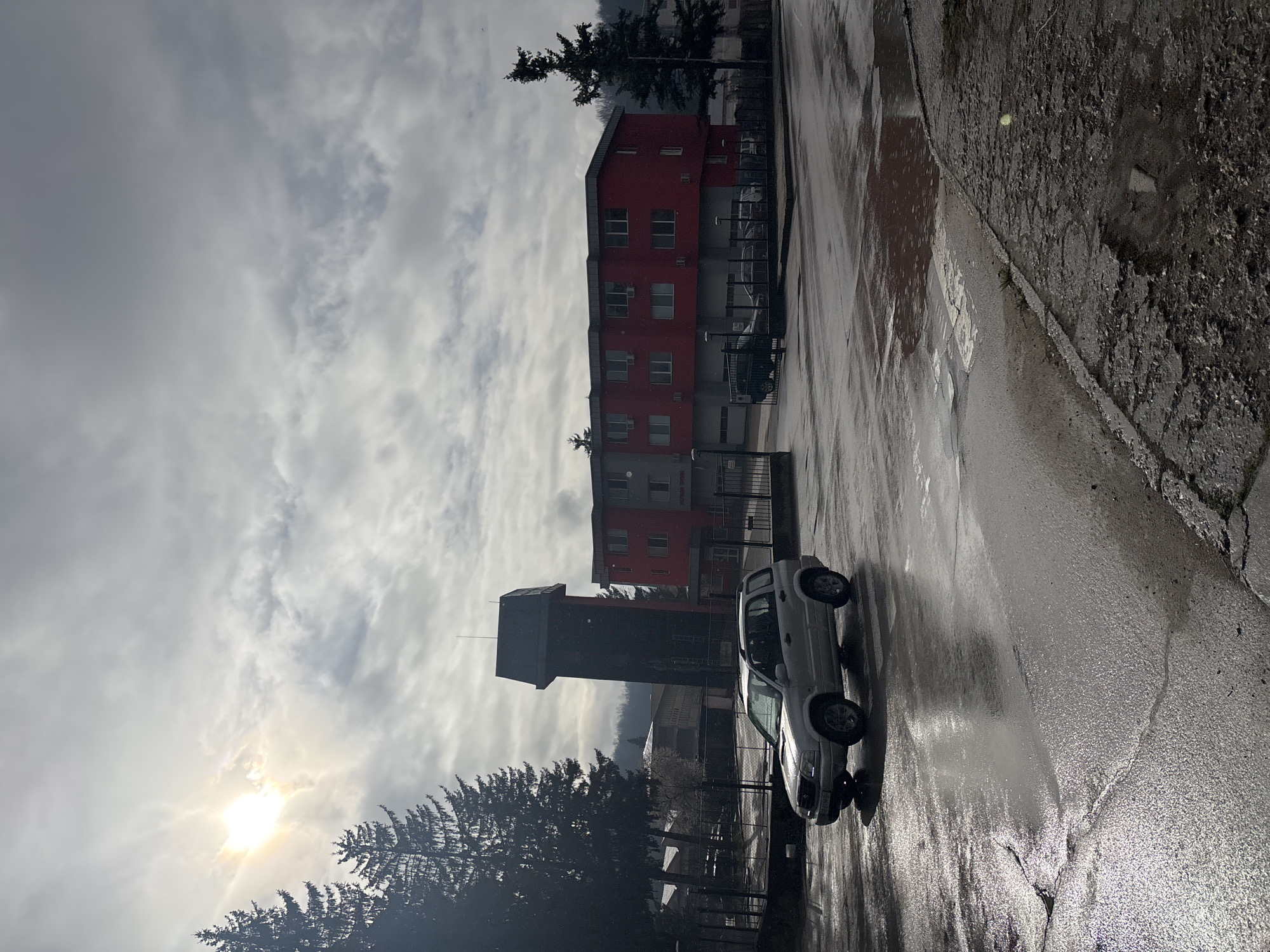 The image captures a street scene in Troyan, Bulgaria, on what appears to be a wet, overcast day. A light-colored pickup truck is parked on the side of the road. Behind the truck stands a tall, dark-colored tower, and a red and grey multistory building. The street is wet, reflecting the sky. The sky is filled with heavy clouds and the sun shines weakly.
