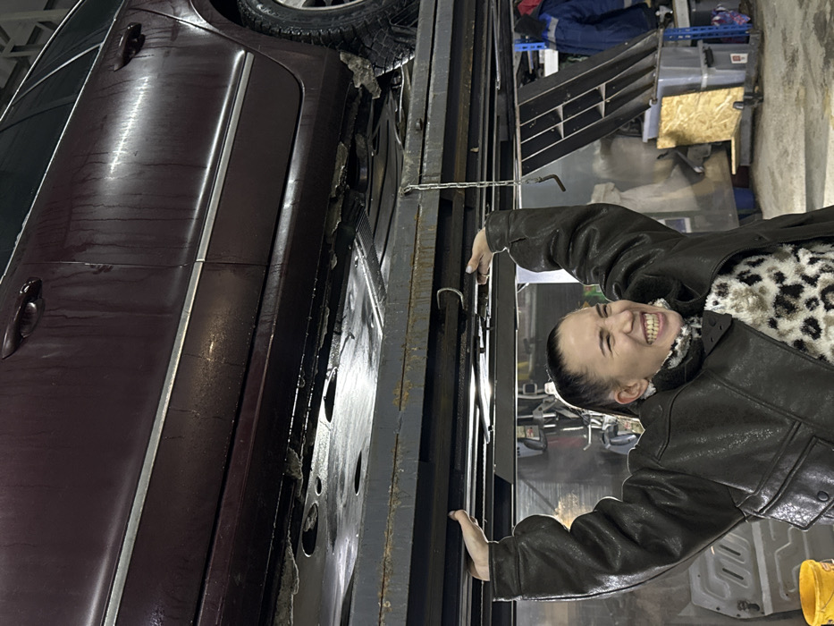 A woman in a brown leather jacket and leopard print sweater stands next to a maroon car on a car lift in a garage. The woman is smiling broadly, her hands on the lift's metal structure. The car is dirty and appears to be undergoing repairs or maintenance. The background includes various garage tools, equipment, and storage shelves. The image is rotated, presenting an unusual perspective.