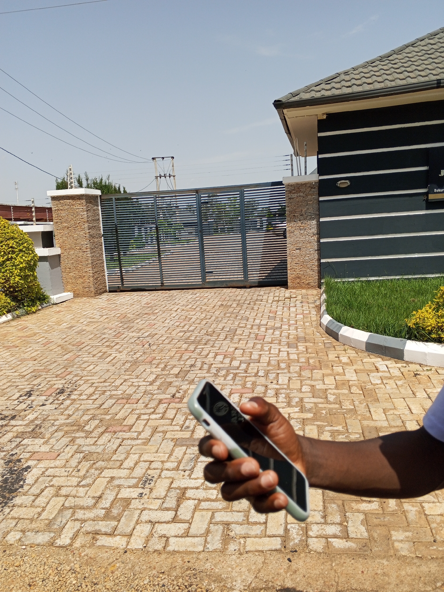 In Bukuru, Nigeria, a person holds a smartphone in their right hand, obscuring a portion of the foreground. The phone, held at an angle, displays a dark screen with a reflection of the surrounding environment. The paved driveway, constructed with interlocking bricks in a herringbone pattern, extends into the middle ground. The driveway is bordered by a neatly manicured lawn and a decorative curb made of white, grey, and black segments. Behind the lawn, a modern building with dark grey siding and horizontal white stripes is visible, featuring a tiled roof. A large, metal gate, likely automated due to its design, stands in the background, supported by brick pillars. Overhead, power lines crisscross the clear blue sky. The scene suggests a residential or commercial property in an urban or suburban setting.