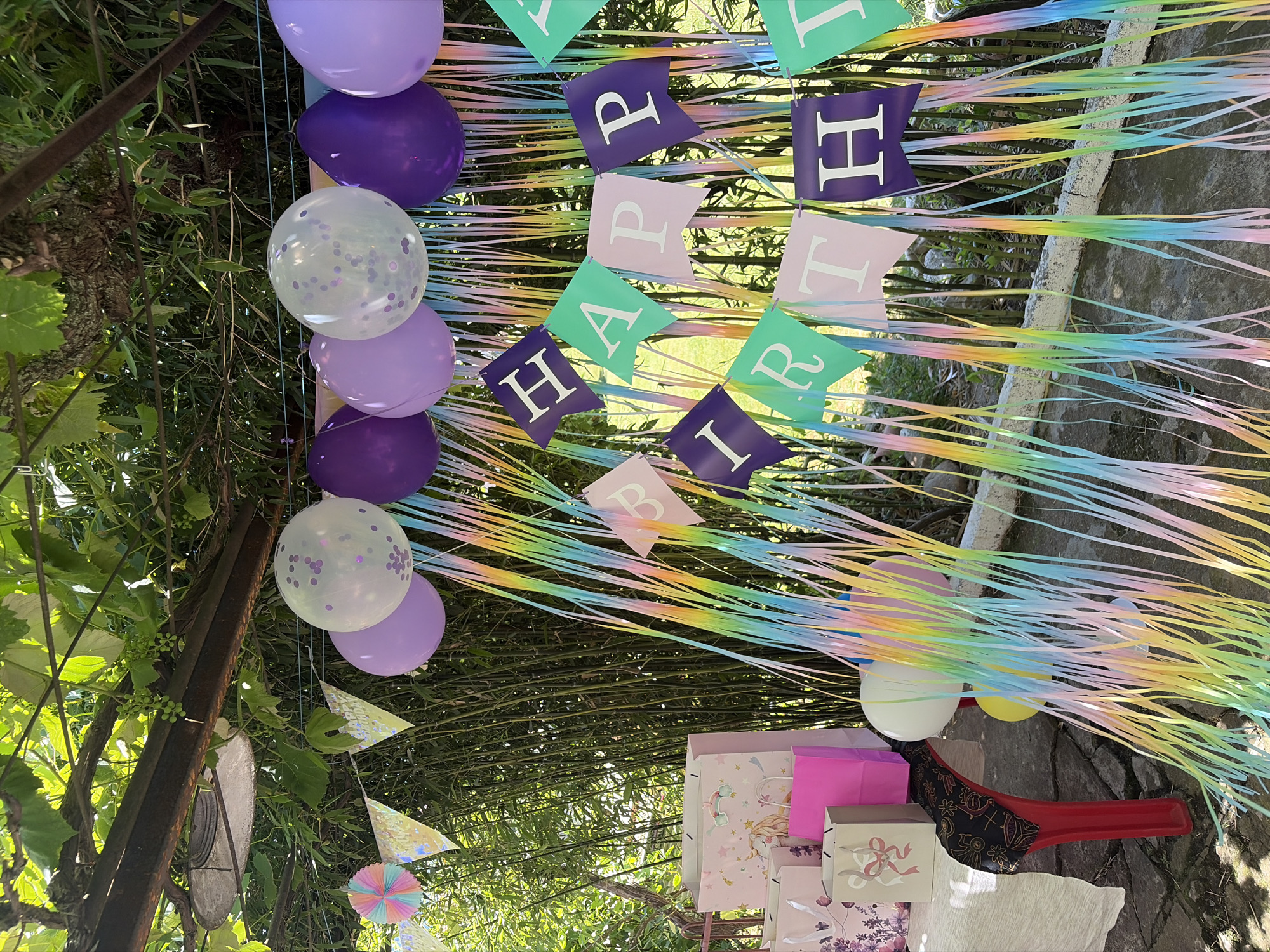 This is a medium shot, eye-level, outdoor photograph of a birthday party setup in Vlasatitsa, Bulgaria. The scene is decorated with balloons, streamers, and banners. Several purple, lavender, and white confetti-filled balloons are suspended from above, interspersed with colorful paper streamers in pastel shades of yellow, pink, blue, green, and purple. A banner with the words "HAPPY BIRTHDAY" spelled out in individual flag pennants of various colors, including purple, pink, and green, is also visible. Gift bags, some decorated with unicorn motifs, are arranged on a table to the left of the frame, along with a red chair with a black, patterned seat. The background consists of lush green bamboo stalks and foliage, suggesting an outdoor, possibly garden, setting. The lighting indicates it is daytime, with sunlight filtering through the leaves above. There are no people present in the photograph.
