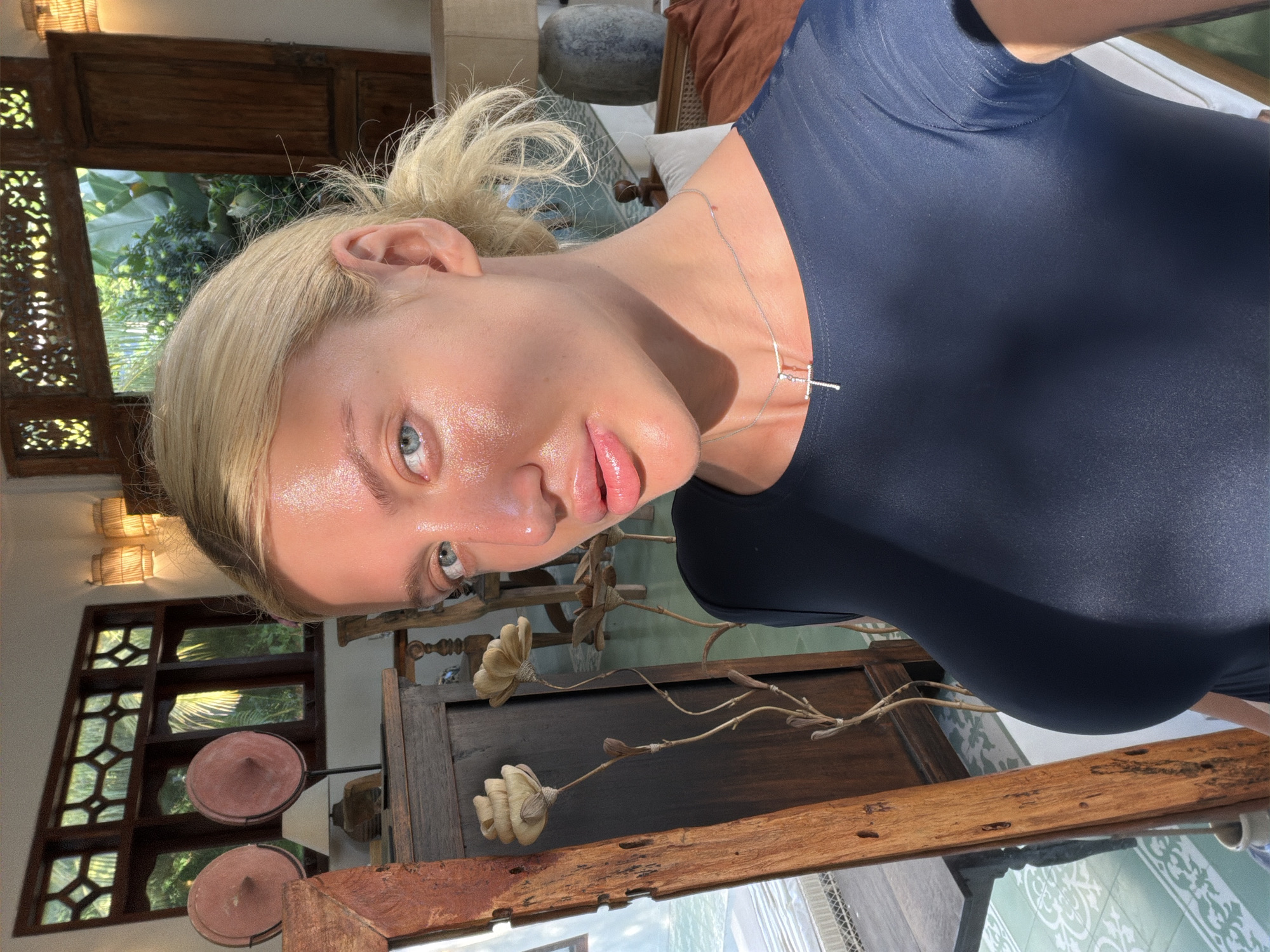In North Kuta, Indonesia, a woman with blonde hair pulled back in a ponytail poses for a selfie indoors. She wears a dark, form-fitting top and a silver necklace with a cross pendant. Sunlight illuminates her face, highlighting her features and causing a prominent sheen on her skin. Behind her, a wooden cabinet with decorative elements stands near a large window with intricate wooden latticework. Two conical decorative objects are placed on the cabinet, and a lamp with a woven shade is visible to the right. The background suggests a room with traditional Indonesian architectural influences, with glimpses of lush greenery visible through the windows.