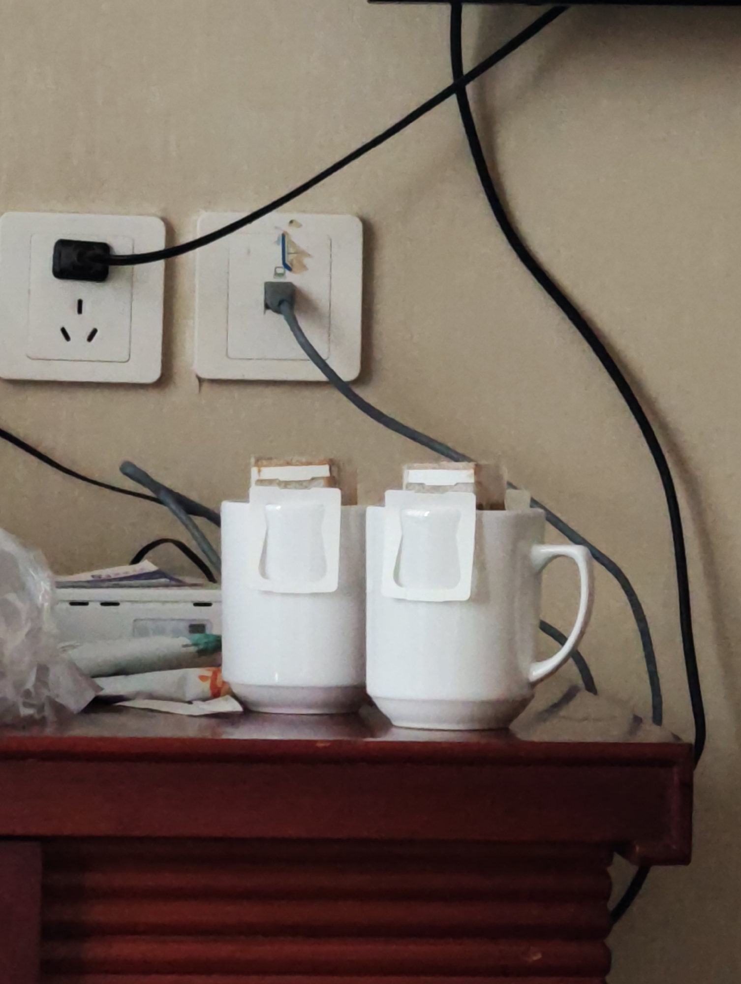 The image shows two white mugs, each with a drip coffee bag hanging over the rim, placed on a dark red wooden surface. Behind the mugs, on a light beige wall, are two white electrical outlets. One outlet has a black plug inserted, and a black cord extends from it, as well as another grey cord. The other outlet has a grey cord inserted. To the left of the mugs, a white electronic device is partially visible, with some packaging or papers in front of it. The overall environment appears to be an indoor setting, possibly a room with a wall and a piece of furniture. The image does not provide any specific details that would allow for precise location within 幸福乡, China.
