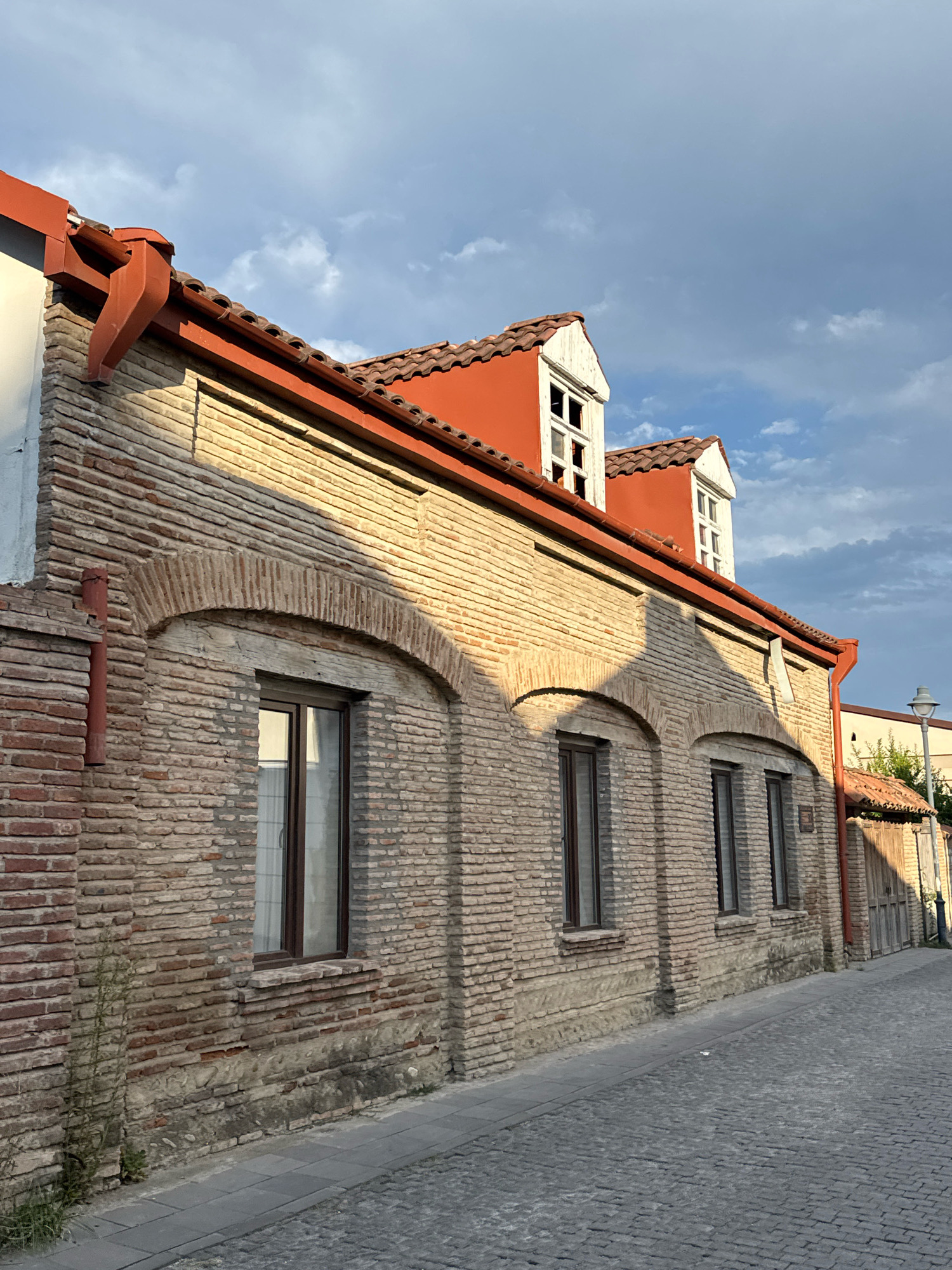 The image depicts a section of an old, two-story building made of light-colored bricks, situated on a cobblestone street in the city of Telavi, Georgia. The building features a red-tiled roof with two dormer windows on the upper level. The lower level has several arched windows with dark frames. Red gutters run along the roofline. The sunlight casts shadows on the brick facade. The street is paved with cobblestones and there is a sidewalk. Vegetation, including grass and a small plant, grows near the base of the building. In the background, a lamppost and a wooden fence are visible. The sky above is partly cloudy with blue sky peeking through. There are no people or animals visible in the image.