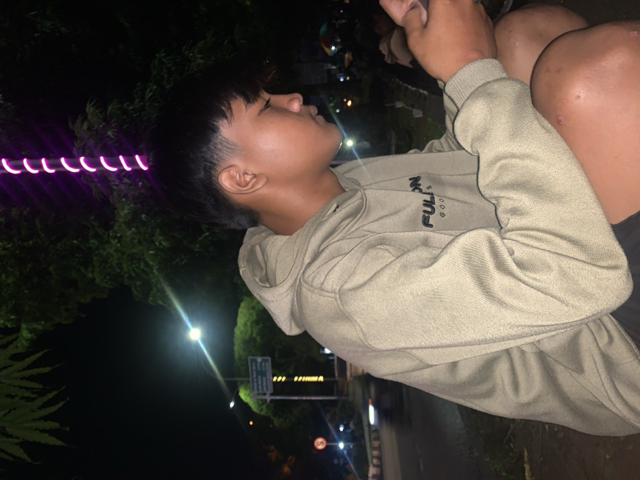 The image presents a side profile of a young individual, likely male, seated outdoors at night in Metro, Indonesia. The person has dark, short hair with a fringe and is wearing a light brown or khaki hooded sweatshirt that features the text "FULON GOODS" in dark lettering on the chest. Their right hand is partially visible, holding an object out of frame, possibly a phone. The individual is looking upwards and to the right with a contemplative or observant expression. Bare knees or legs are visible on the right side of the frame, suggesting a relaxed, seated posture.
The background is dark, indicating nighttime, illuminated by various artificial light sources. A distinctive purple, spiraling light trail is prominent in the upper left of the frame. Bright streetlights or overhead lamps create flares and cast light on the surrounding environment, which includes visible trees and foliage. A road or pathway stretches into the distance, with a dimly visible sign featuring a "5" inside a circle, potentially a speed limit indicator, and another unreadable road sign further along. The scene conveys a quiet, nocturnal urban or semi-urban atmosphere.