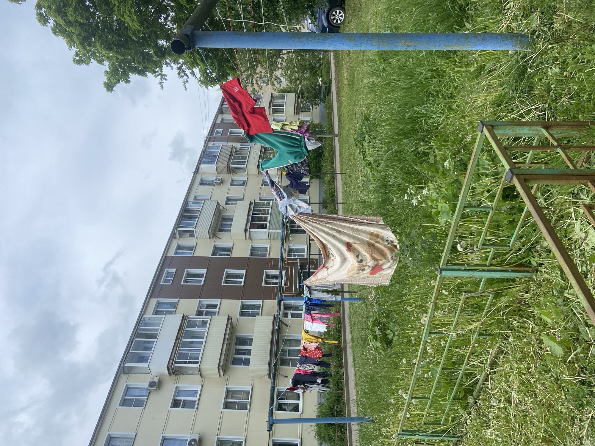 The image depicts a clothesline strung between two blue metal posts in Peresvet, Russia. A variety of clothing items are hanging on the line, including a red shirt, green sweatpants, and a blanket with a teddy bear pattern. A multi-story apartment building with beige and brown paneling is visible in the background. The ground is covered in green grass and weeds. A green metal frame of what may be a swing set or other playground equipment is situated to the right of the clothesline. The sky is overcast.