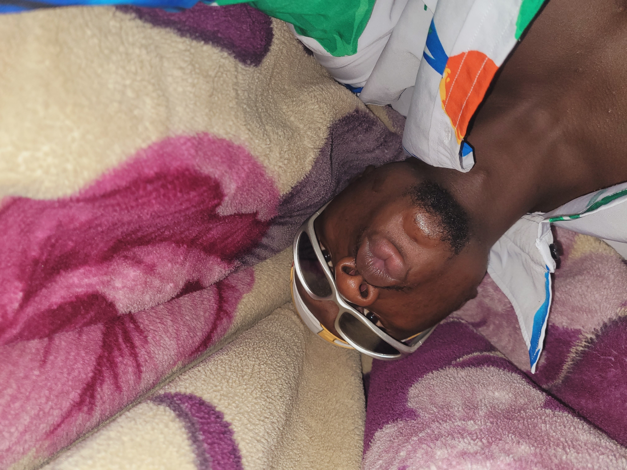 The image shows a close-up, head-and-shoulders view of a dark-skinned male lying down, possibly in bed. He is wearing white sunglasses with a silver frame and a white shirt with a colorful pattern that includes orange, green, and blue shapes. His lips are slightly pursed. He has a short beard. The background consists of a textured blanket with a floral pattern in shades of purple, pink, and beige. The image appears to have been taken indoors. The location in Bukuru, Nigeria, is not specified within the image itself and cannot be inferred from the visual information provided.