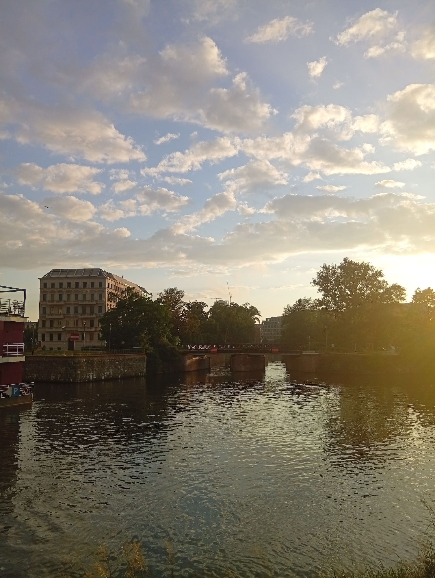 The image depicts a wide river in Wrocław, Poland, flowing from right to left. On the left bank, a multi-story building with numerous windows and a flat roof stands prominently. Adjacent to it is a red structure with white railings, possibly a docked boat or a pier. The river is crossed by a bridge, visible in the middle ground, with brick pillars supporting its structure. The right bank is lined with trees, some of which are large and mature. The sky is filled with scattered white clouds against a blue background, with the setting sun casting a warm glow on the right side of the frame. The water reflects the sky and the surrounding environment. There are no visible people in the image.