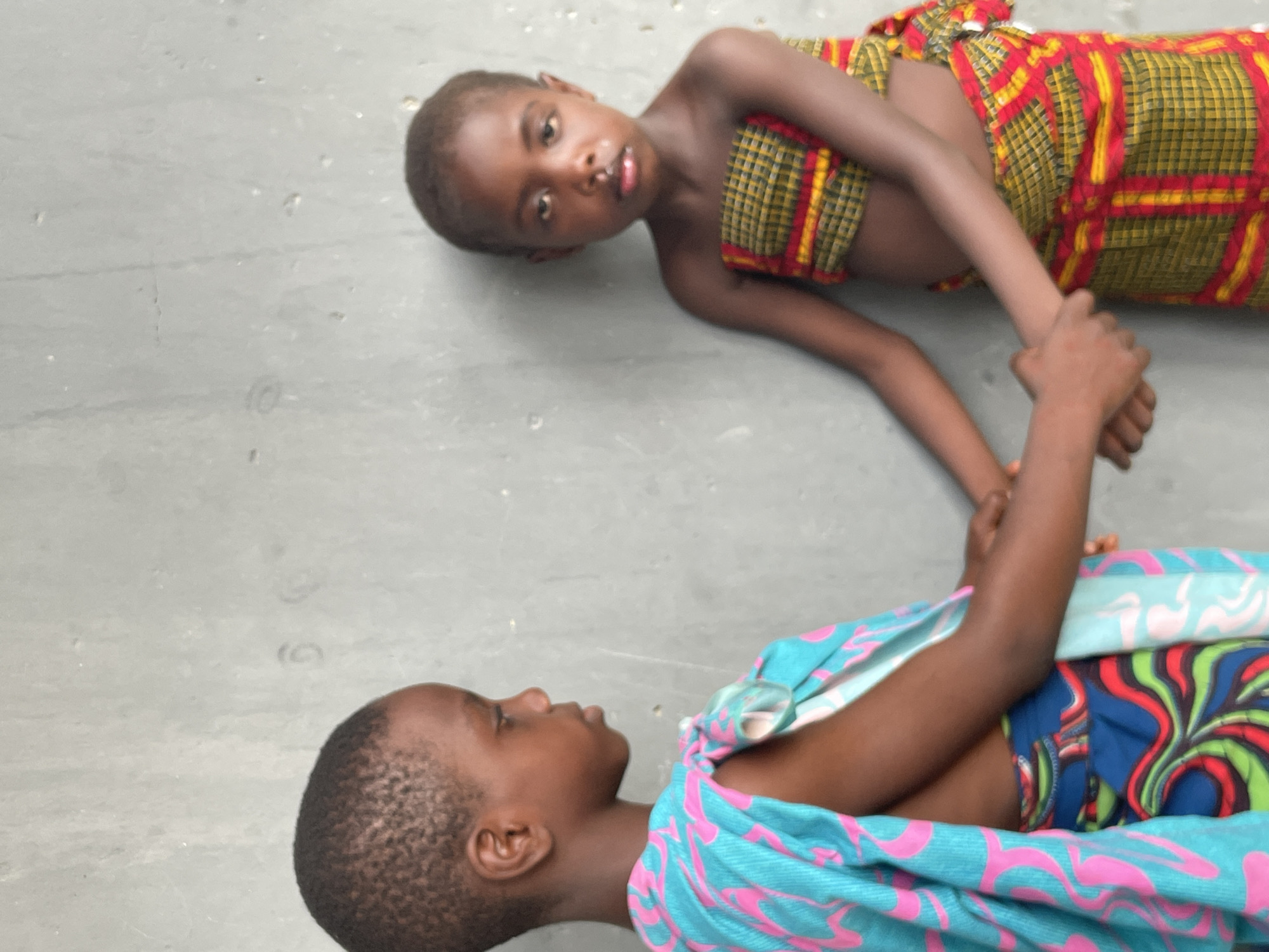 Two young children are lying on a light grey, possibly concrete or paved, surface in Badagry, Nigeria. They are positioned head-to-head, facing opposite directions, with their hands clasped together in the center of the frame.
The child on the upper left side of the image has dark skin and short, dark hair. They are wearing a fabric wrap with a prominent red, yellow, and black plaid or checkered pattern across their chest and lower body. This child is looking directly at the camera with a neutral expression.
The second child, on the lower right, also has dark skin and short, dark hair. They are dressed in a brightly colored fabric wrap, predominantly blue with swirling pink patterns, over another garment visible underneath featuring blue, red, green, and black abstract designs. This child is seen in profile, looking towards the first child or slightly beyond, with a thoughtful or serious expression.
The setting appears to be outdoors during the daytime, characterized by bright and even lighting, suggesting clear weather. The simple, light-colored ground provides a neutral backdrop, highlighting the children and their colorful attire. There is no visible text in the image. The scene captures a moment of connection and perhaps play between the two children.