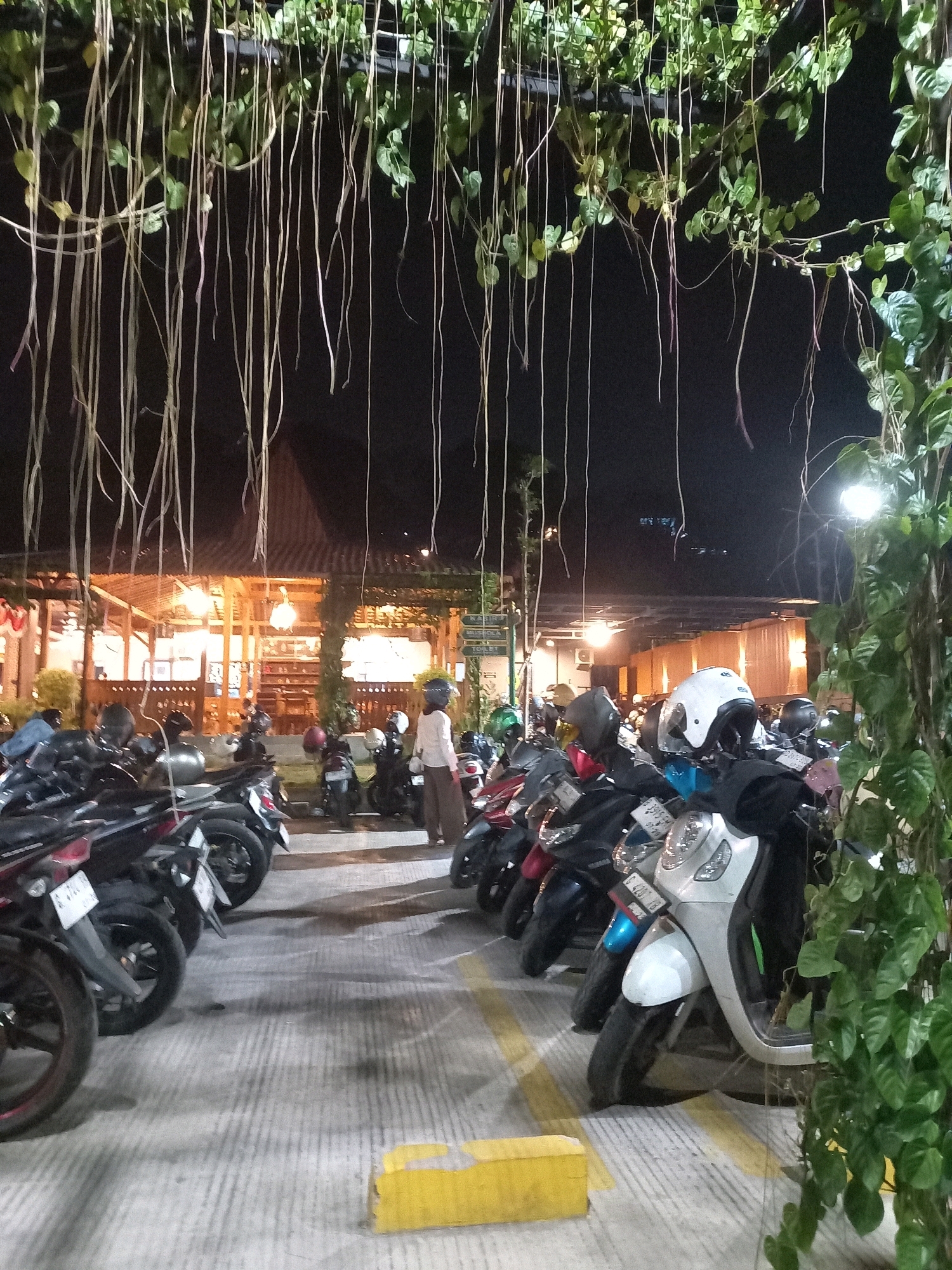 This is a nighttime photograph of a parking lot filled with parked scooters and motorcycles in front of a dimly lit, open-air establishment with a traditional Indonesian-style roof. The establishment appears to be a restaurant or food stall, indicated by the warm lighting and tables visible inside. Hanging vines from the overhead structure obscure parts of the scene, creating a somewhat enclosed atmosphere. Several people are visible, including a person in a white helmet standing near a row of motorcycles and another person walking in the background. The ground is paved with concrete and marked with yellow lines. A street lamp provides illumination in the upper right portion of the image. The overall scene suggests a casual, social gathering spot that is popular in the evening. The presence of the specific license plate format "S 4265 YB" on one of the scooters indicates the location is likely in the East Java region of Indonesia, though the request specifically asks for Semarang. Given the ambiguity of the license plate and the request for Semarang, this could be a provincial tourist spot or a place that attracts visitors from various regions.