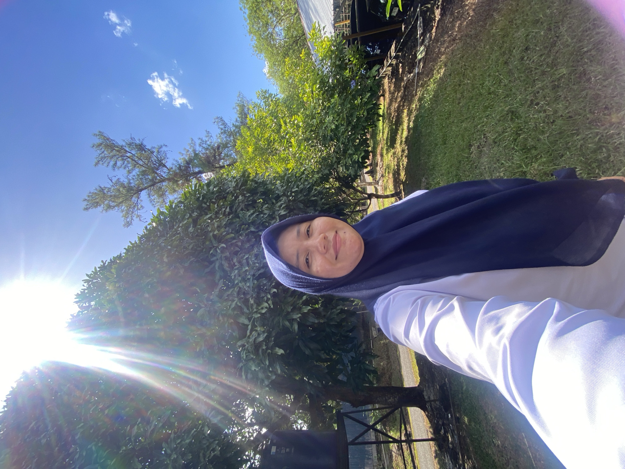 A young woman in a dark blue hijab and a white shirt is taking a selfie outdoors. She is smiling at the camera, and the sun is shining brightly from the upper left corner of the image, casting lens flares. Behind her, there are trees with lush green leaves and a clear blue sky with a few white clouds. In the background, a large black water tank on a metal stand is visible to the left, and to the right, there appears to be a covered structure with a metal roof. The ground is covered with grass and some dirt paths. The scene suggests a park or a natural area within the city of Banjarbaru, Indonesia, possibly a recreational space or an educational institution.