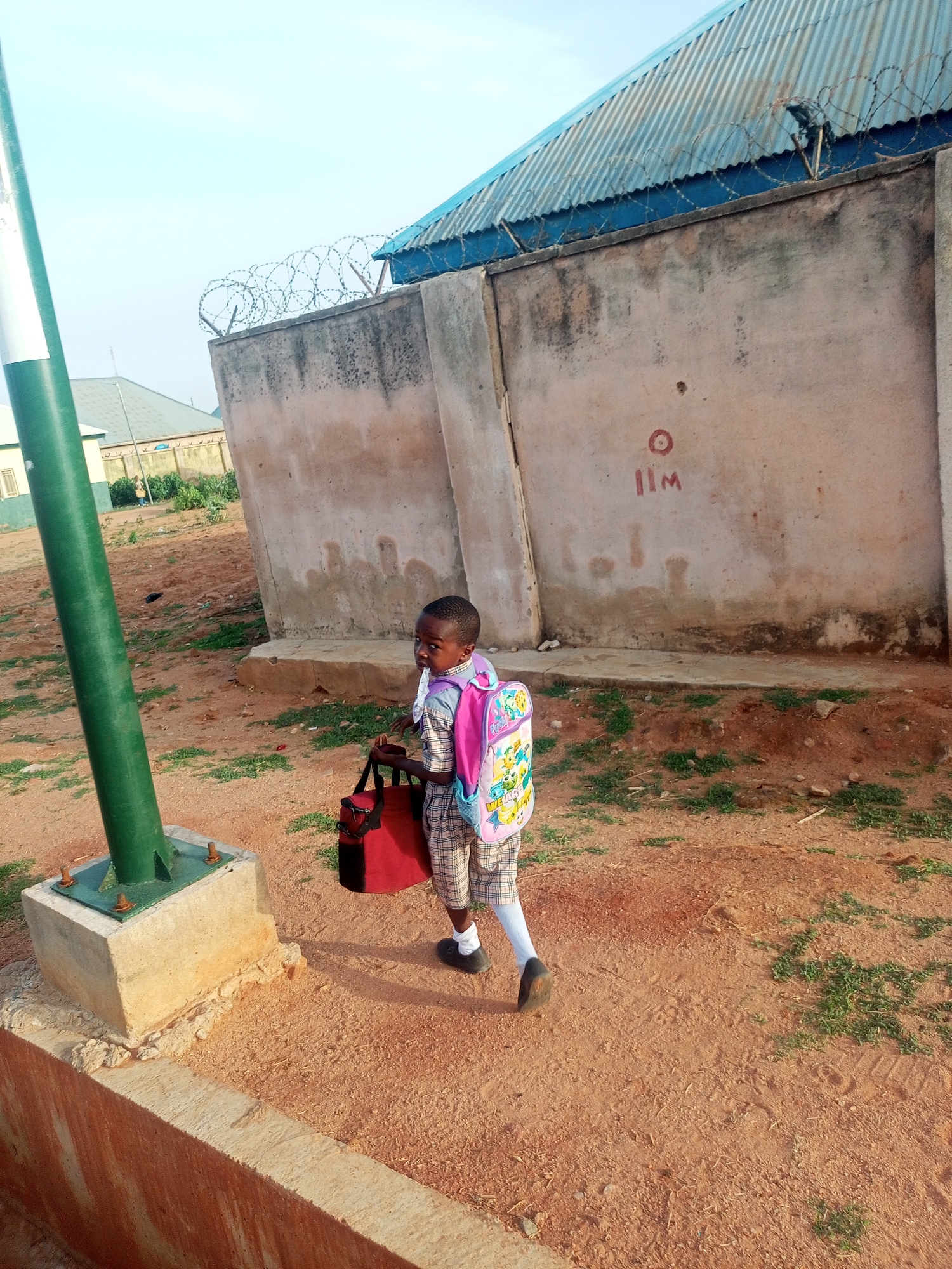 A young boy is walking on a dirt and grass-covered ground in what appears to be an outdoor area near a building. He is carrying a red bag in his left hand and has a pink backpack on his back. The boy is wearing a plaid shirt, plaid shorts, white socks, and dark shoes. Behind him is a tall concrete wall topped with barbed wire. The wall is discolored with patches and writing on it. To the left of the boy is a tall, green and white pole set in a concrete base. In the background, there is another building with a light blue corrugated iron roof. The scene seems to be situated in Tempure, Nigeria. The time of day appears to be early, given the soft lighting and shadows.
