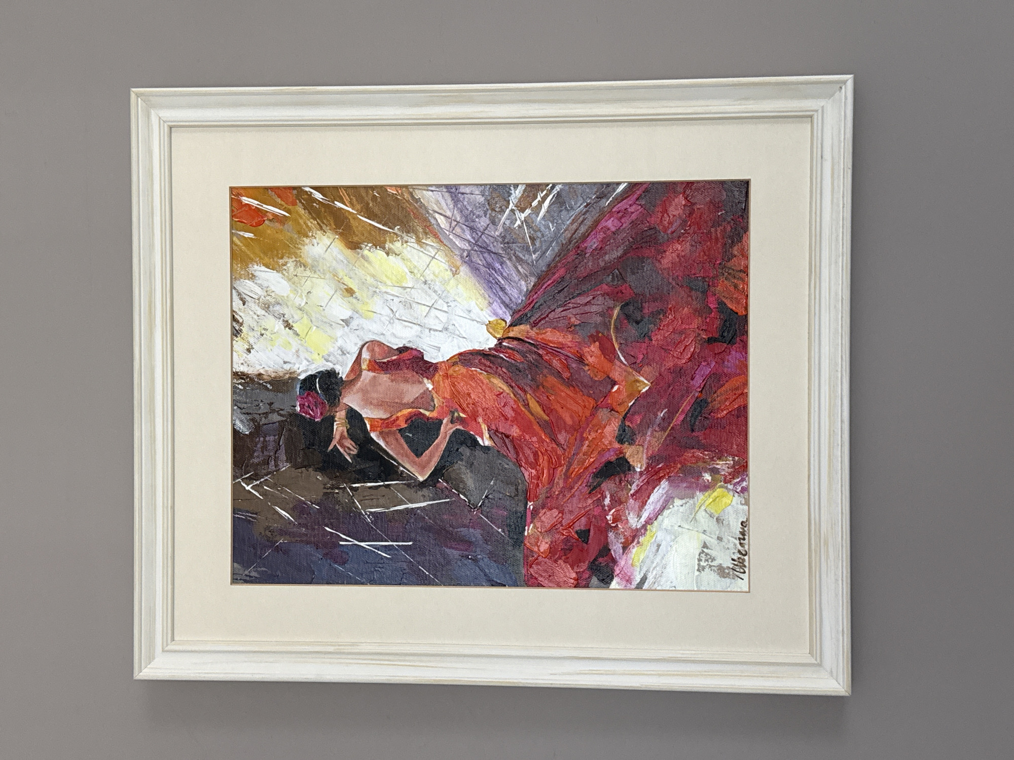 A framed painting of a flamenco dancer is displayed on a gray wall. The painting depicts a woman in a vibrant red flamenco dress, her back to the viewer, with her arms outstretched and her body in a dynamic pose. The background is abstract and energetic, with splashes of yellow, brown, and white, suggesting movement and passion. The dancer has a red flower in her hair and wears gold bracelets on her wrists. The signature of the artist, "Milena", is visible in the bottom left corner of the painting. The artwork is framed with a white, textured mat and a distressed white wooden frame. This painting is part of the collection displayed at the "Dimitar Dobrevich" Municipal Art Gallery, located at 4 Ul. Bogoridi, Burgas, Bulgaria.