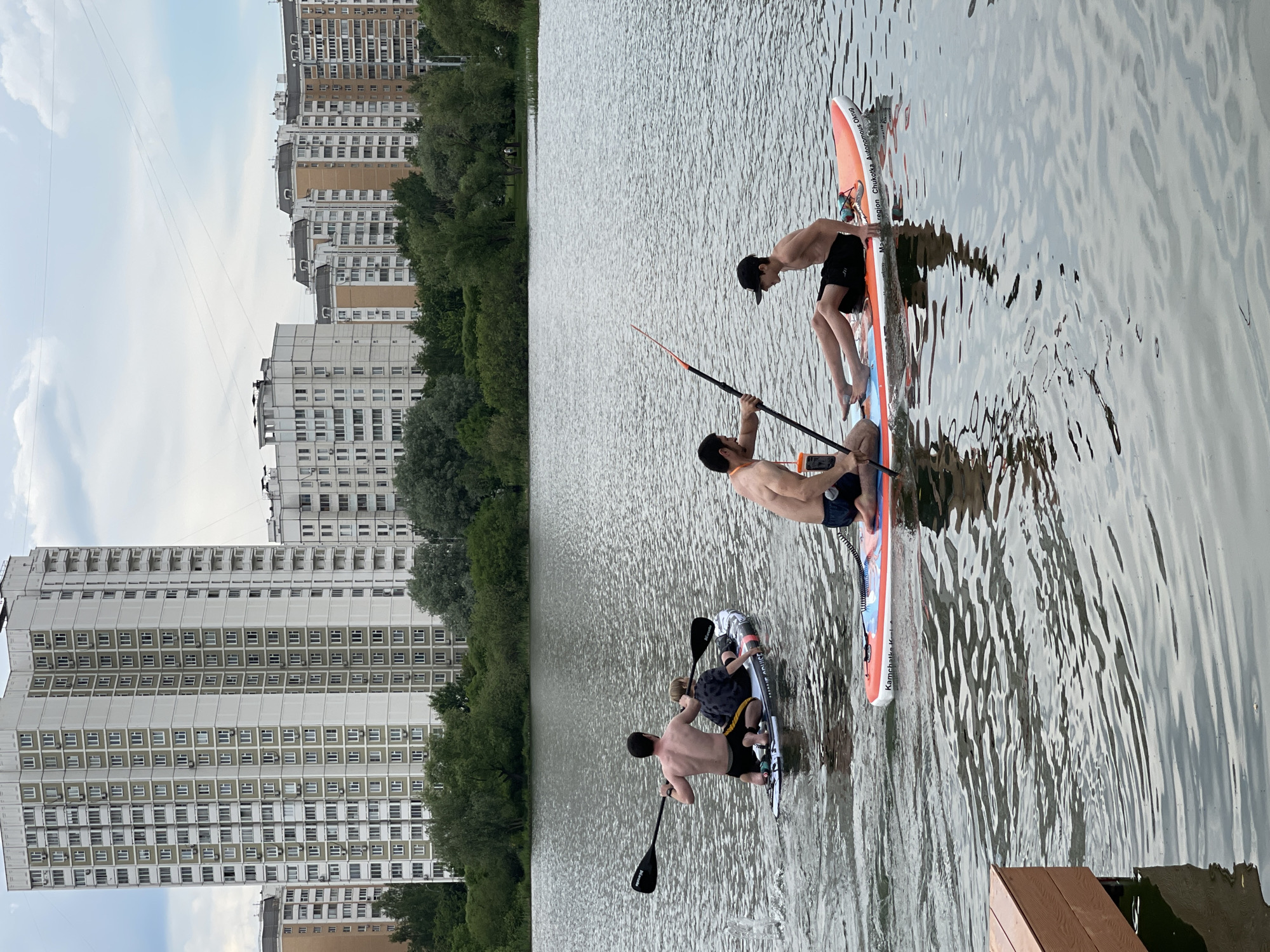 Three individuals are engaged in paddleboarding on a calm body of water, likely a lake or river, under an overcast sky. In the foreground, two men, shirtless and wearing swim shorts, are actively paddling on separate stand-up paddleboards (SUPs). One man is kneeling on a grey and black SUP, paddling with a black paddle, while another man is on an orange and white SUP, also holding a paddle. A younger person, wearing a black cap and dark shorts, is seated on the front of the orange and white SUP, facing the first man.
The background features a densely built urban environment, with multiple tall, multi-story residential apartment buildings visible through a line of green trees that separates the waterway from the city. The buildings are primarily light-colored, suggesting typical post-Soviet era architecture, consistent with the given location context of Moscow, Russia. The sky is largely white and cloudy, indicating an overcast day.
Notable details include text visible on the paddleboards: The orange and white SUP has "BIKKO smolbolius Autono" and "Champion Chikoka Region" printed on its side. Another board, partially visible on the far left, shows text like "Kamchatka" and "K-S". A small wooden dock or platform is visible in the bottom right corner of the frame. The water surface shows gentle ripples, reflecting the subdued light. The overall scene depicts a leisurely recreational activity in an urban park or waterfront area.