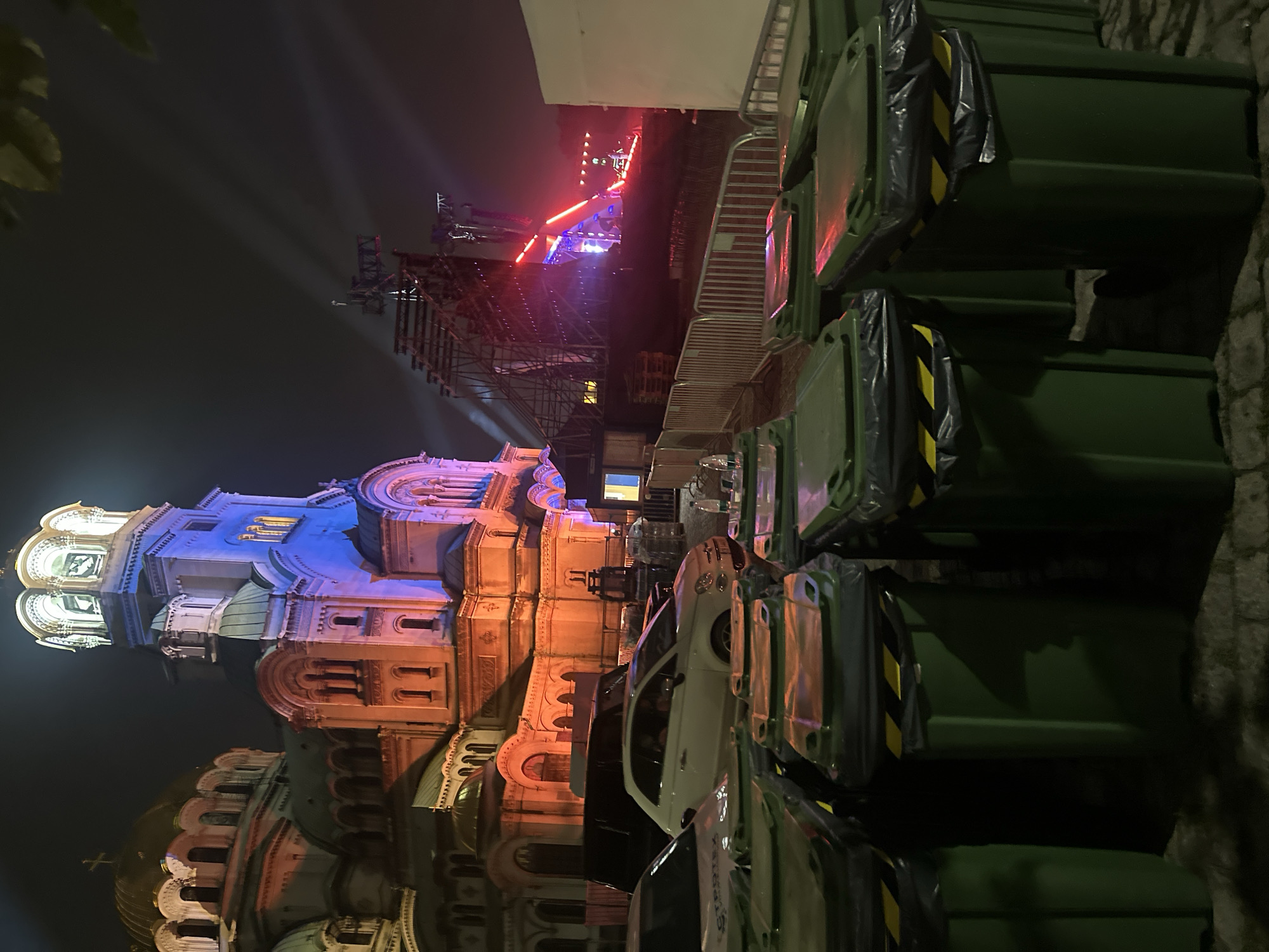 The image shows a large, ornate building illuminated with orange and blue lights at night. In the foreground, a row of dark green trash bins with yellow and black hazard tape is visible. Behind the bins, several cars are parked, including a white Fiat 500. Further in the background, a large stage structure with bright lights and scaffolding is set up. The overall scene suggests an outdoor event or concert taking place near a historical building. The location is not specified.