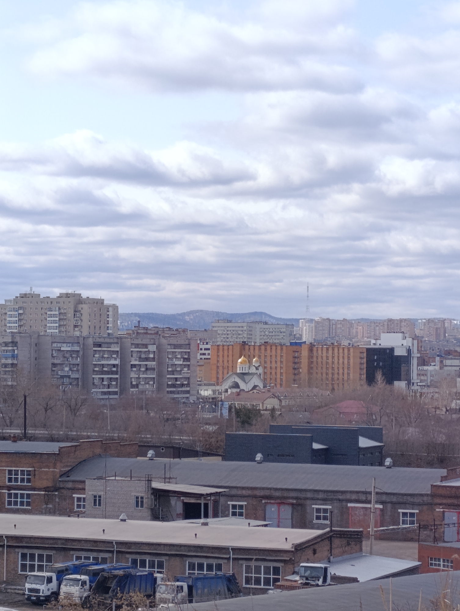 The image depicts a cityscape in Krasnoyarsk, Russia, under a cloudy sky. In the foreground are low-rise industrial buildings, constructed primarily of brick and concrete, with flat or gently sloping roofs. Several trucks are parked near one of the buildings. In the middle ground, there are residential buildings, including apartment blocks of varying heights and colors, with one prominent building having orange facades. A church with golden domes is visible among the residential buildings. In the background, there are mountains or hills on the horizon, slightly obscured by haze. A transmission tower is located among the buildings on the horizon. The overall atmosphere is urban and somewhat industrial, with a mix of residential and commercial architecture. The season appears to be late autumn or early spring, given the bare trees and overcast sky.