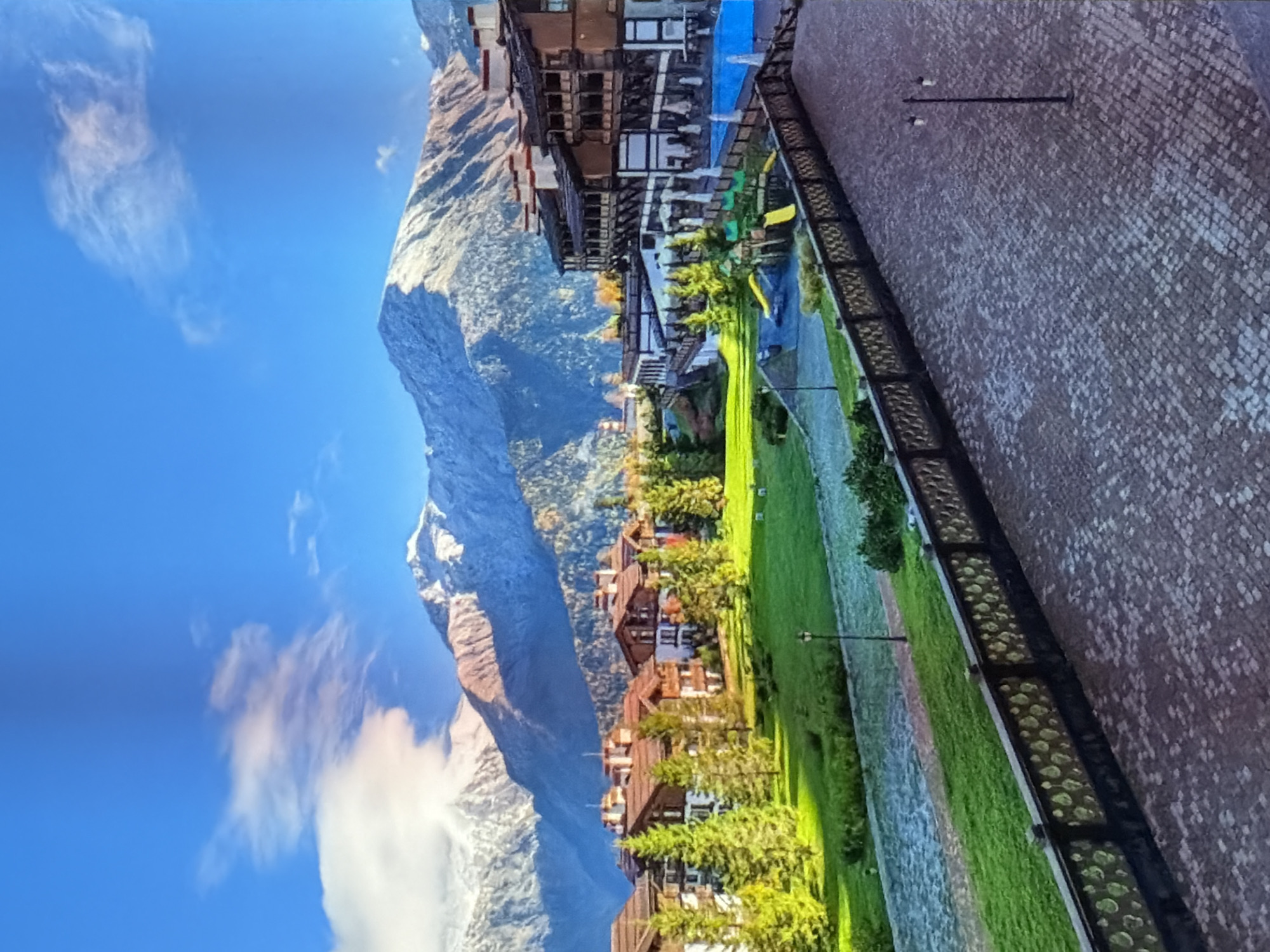 The image displays a landscape with a mountain range to the left and a series of buildings and green space to the right. The mountain has a mix of rocky and snowy terrain with clouds near the base. The buildings, likely residences, appear to be multi-storied structures with dark roofs. A patch of green space separates the buildings from a paved area on the right, which seems to be a brick or cobblestone pathway, possibly a sidewalk or plaza. There are evenly spaced street lamps along the pavement, indicating an urban or developed setting in an unknown location.