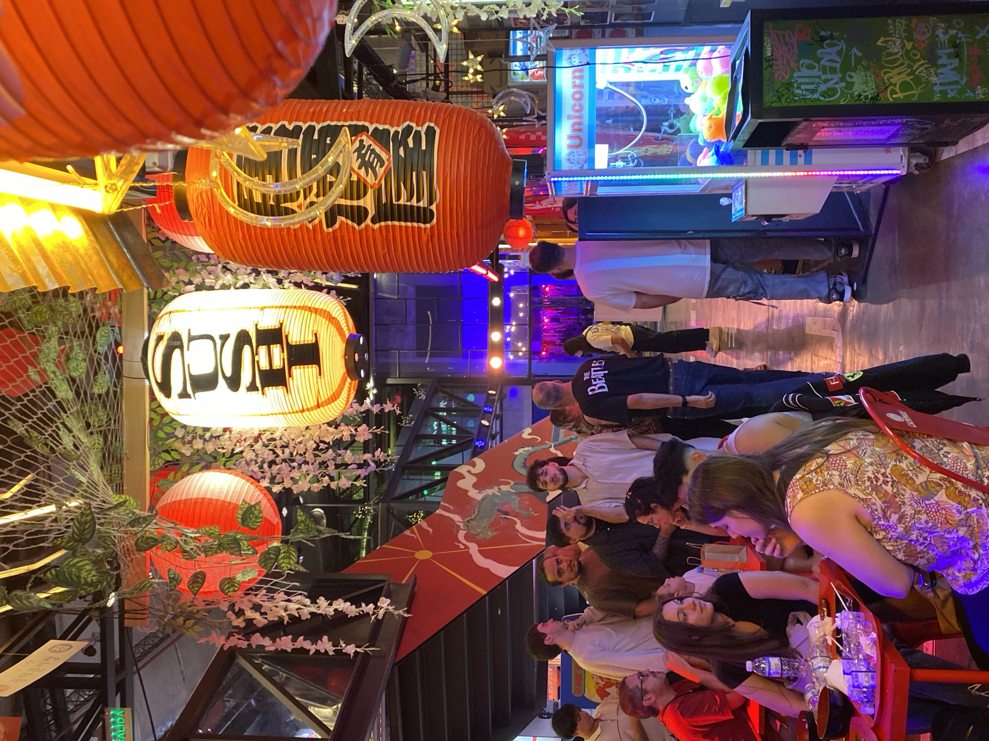Here is a forensic description of the image:
**Setting:** Indoor establishment, likely a restaurant or bar, with East Asian decor.
**Decor:** Prominent hanging paper lanterns in red, orange, and white with East Asian characters. There are additional hanging decorations, including artificial plants and twinkling lights. A wall mural depicting a dragon and the sun is visible.
**Objects:** Tables and red chairs are set up. A claw machine arcade game stands along a wall. Bottled water and glasses are on the tables.
**People:** A group of at least a dozen people are seated at the tables. Some are interacting with each other, appearing to be engaged in conversation. Individuals of various ages and genders are present. Clothing is casual. Two people are standing in the background playing the claw machine game.
**Location:** The original prompt stated this image takes place in Buenos Aires, Argentina.
