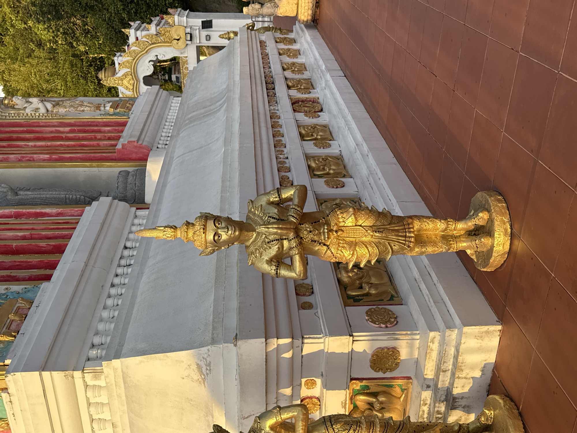 The image shows a close-up view of a golden statue of a male figure in a traditional Thai stance with hands pressed together in prayer, positioned in front of a white temple structure. The statue is adorned with intricate details, including a crown, jewelry, and ornate clothing. The temple features a white tiered base with a decorative frieze of golden relief figures and a balustrade with small pillars. Behind the statue, the temple wall is painted with red and gold vertical stripes. To the right, a lush green hillside with trees is visible, indicating an outdoor setting. The ground is paved with reddish-brown square tiles. The lighting suggests it is late afternoon, with warm sunlight casting long shadows. The statue is a focal point, with another partially visible golden statue to its left and another smaller statue on a pedestal in the background to the right. The overall scene conveys a sense of religious or cultural significance within a temple complex, possibly located in Patong, Thailand, given the common presence of such religious art in the area.