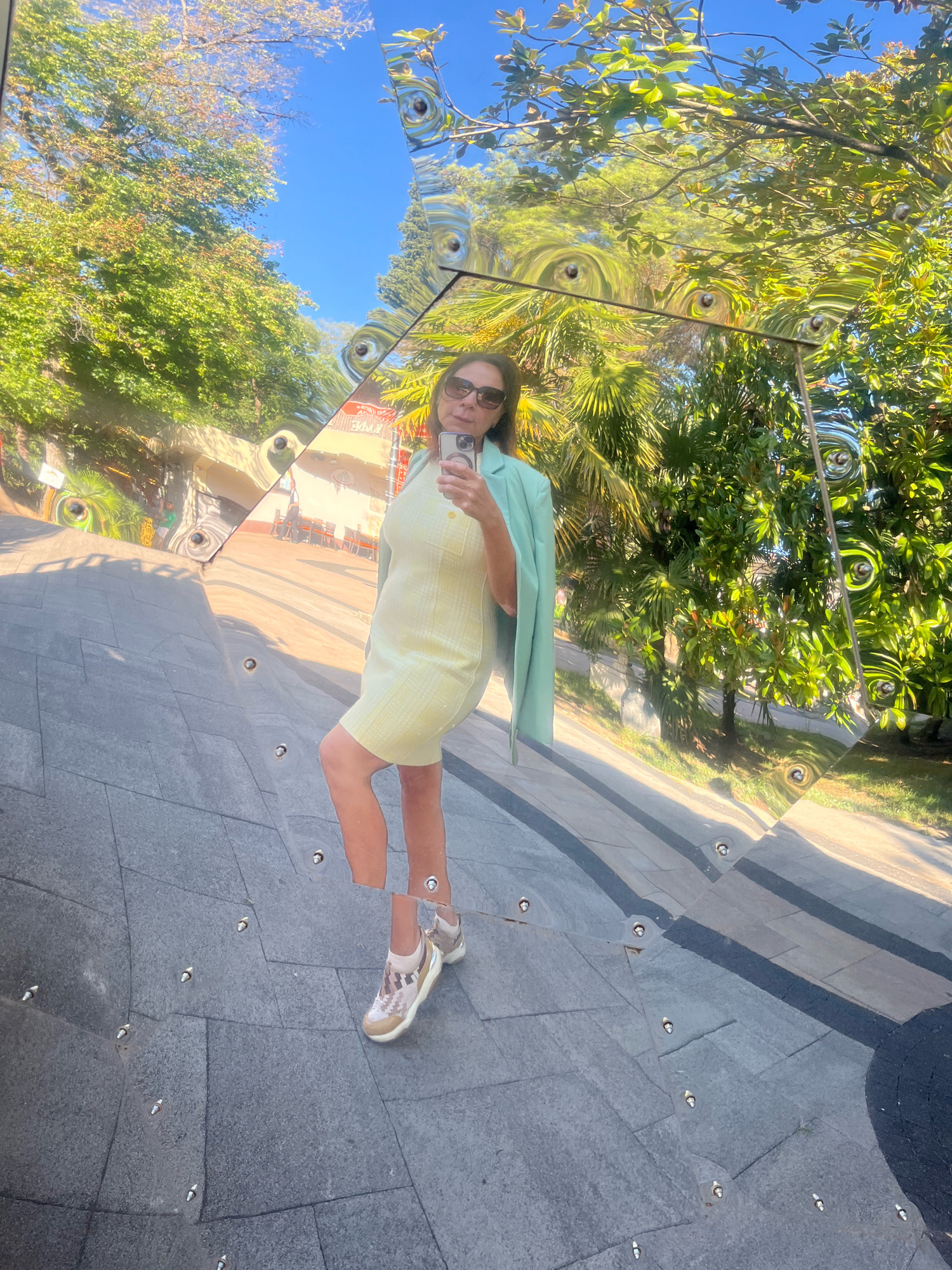 A woman stands in front of a large, abstract, reflective sculpture in Sochi, Russia. She is wearing a pale yellow ribbed dress, a light mint green blazer draped over her shoulders, sunglasses, and light-colored sneakers. She is holding a smartphone in her right hand and taking a selfie. The sculpture reflects the surrounding environment, including trees, a blue sky, and parts of a street or pathway with people and tables in the background. The sculpture is made of polished metal and has a distorted, warped reflection. The ground beneath the sculpture is paved with gray stones. The image captures a sunny day with lush green foliage.