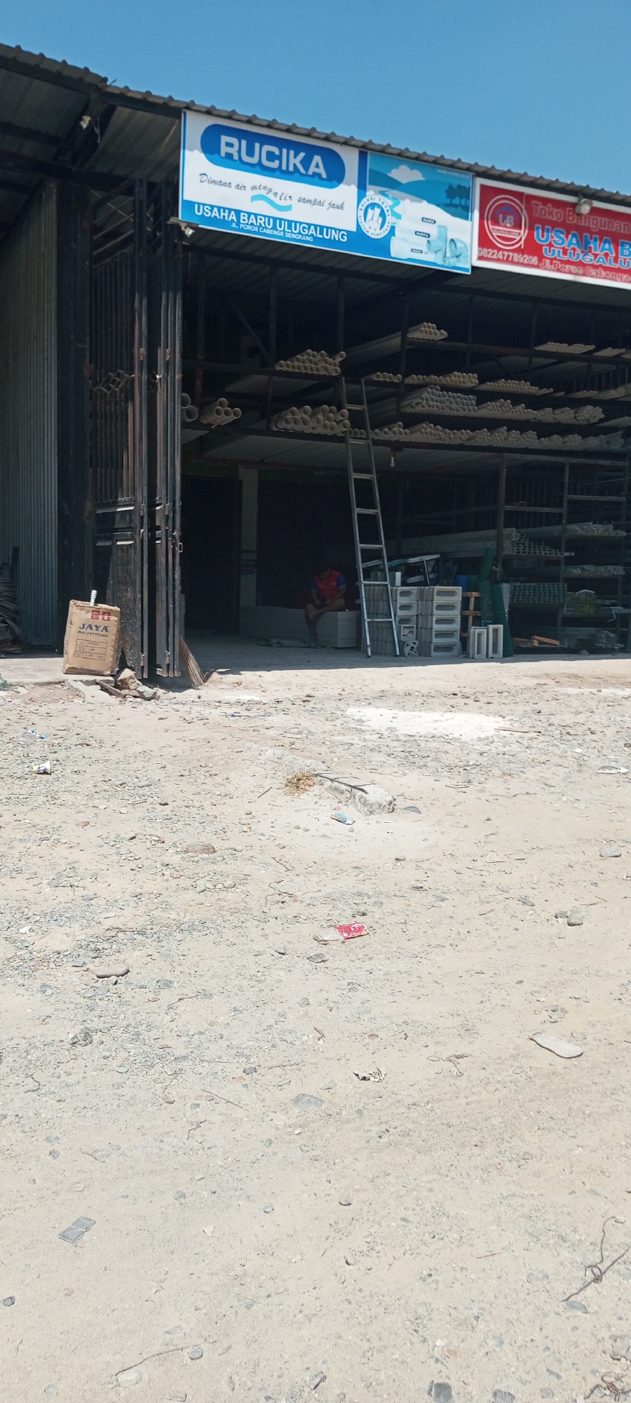 The image shows the exterior of a building with a metal roof, likely a store or warehouse, under a clear blue sky. A sign above the entrance reads "RUCIA" and below it, "USAHA BARU ULUGALUNG" with an address likely in Lempa, Indonesia. Several shelves inside are filled with pipes and building materials. A ladder stands against one of the shelves. In the foreground, the ground is rough, composed of dirt, gravel, and some scattered debris. A person is seated inside the building.