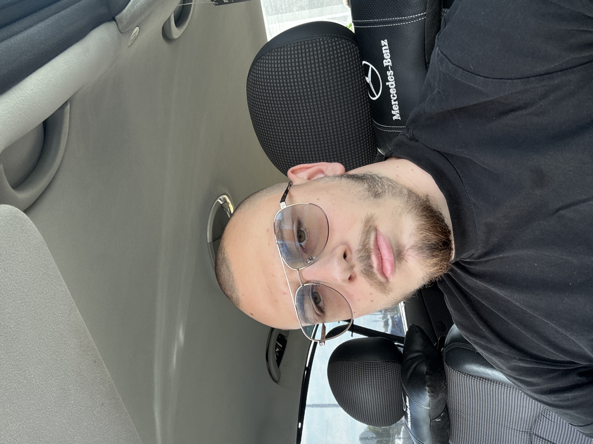 A man is in a vehicle, likely a car, due to the presence of a headrest that has "Mercedes-Benz" written on it. The man is fair-skinned and has a beard and a shaved head. He is wearing glasses and a black shirt. The car's interior is light gray. The location is unknown, but the prompt mentions Stara Zagora, Bulgaria.