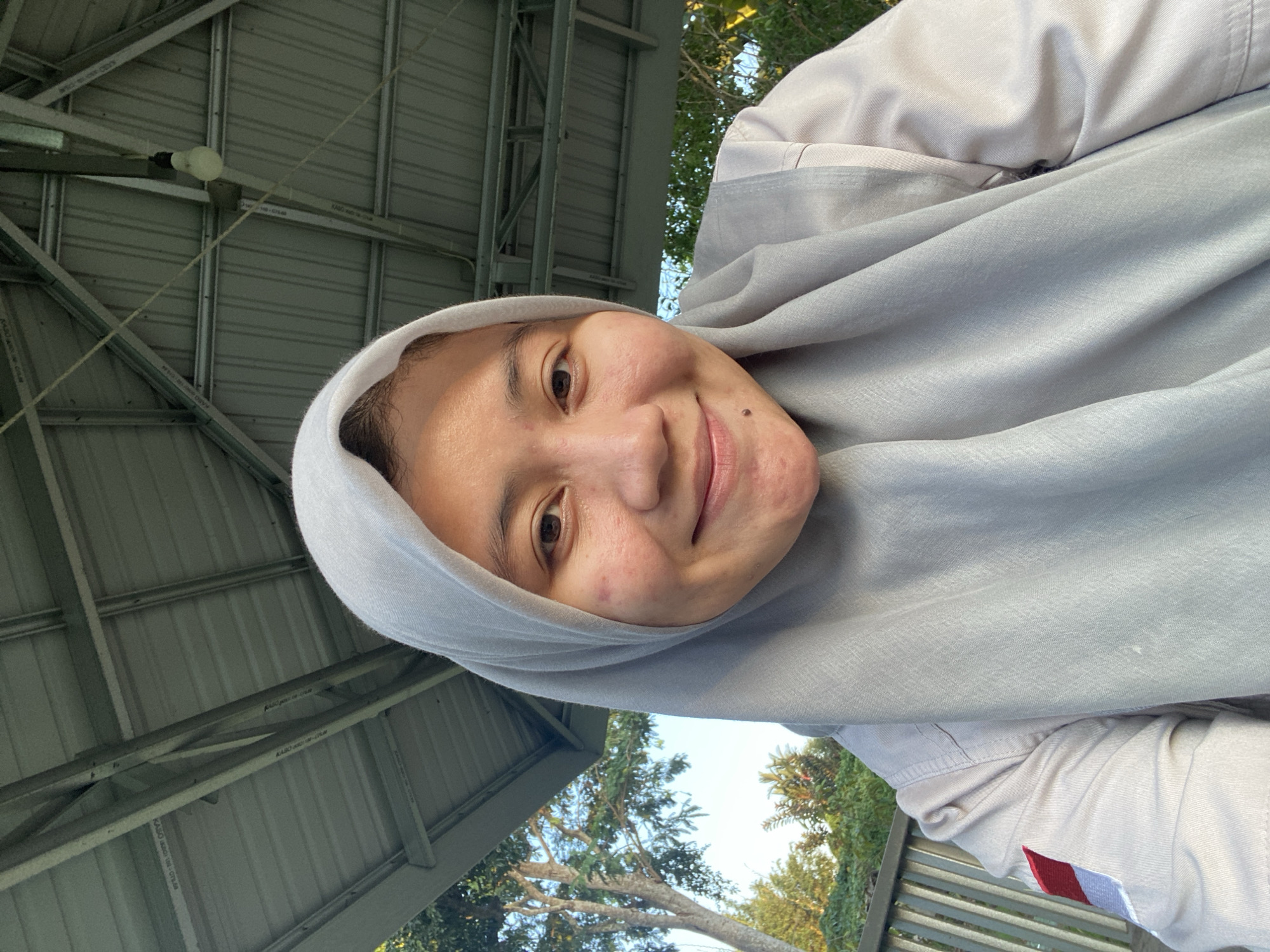 A young woman wearing a light grey hijab and a light-colored collared shirt is pictured in a close-up, selfie-style shot. She has a mole on her chin and some acne on her cheeks. She is smiling. She appears to be outdoors under a metal roofed structure, with trees and foliage visible in the background. A red and white patch is visible on the sleeve of her shirt. The image was taken in Banjarbaru, Indonesia.