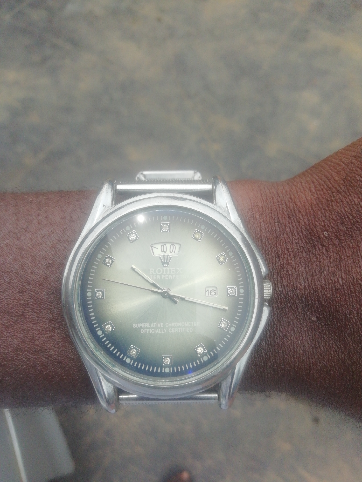 A close-up, eye-level photograph shows a dark-skinned arm wearing a silver-toned wristwatch, centered in the frame. The background is out of focus, appearing as a blurred mix of light grey and brownish tones, possibly concrete or pavement, suggesting an outdoor setting.
The watch features a round, polished silver-colored case with matching lugs and a partial view of its metal bracelet. The watch face is a greenish-grey color with a sunburst effect. Most hour markers are represented by small, square-cut, clear gemstone-like accents. At the 12 o'clock position, there is a crown logo followed by the visible text "ROLEX" and "OYSTER PERPETUAL". A date window at the 3 o'clock position displays the number "16". Above the 6 o'clock position, the words "SUPERLATIVE CHRONOMETER OFFICIALLY CERTIFIED" are inscribed on the dial. The watch has three hands: hour, minute, and a sweeping second hand. Significant reflections and glare are visible across the watch's crystal, particularly in the upper left and lower right quadrants. The skin on the arm is visibly textured with fine hair.
The image captures the static display of the watch on the wrist. There are no discernible activities or specific time of day cues.