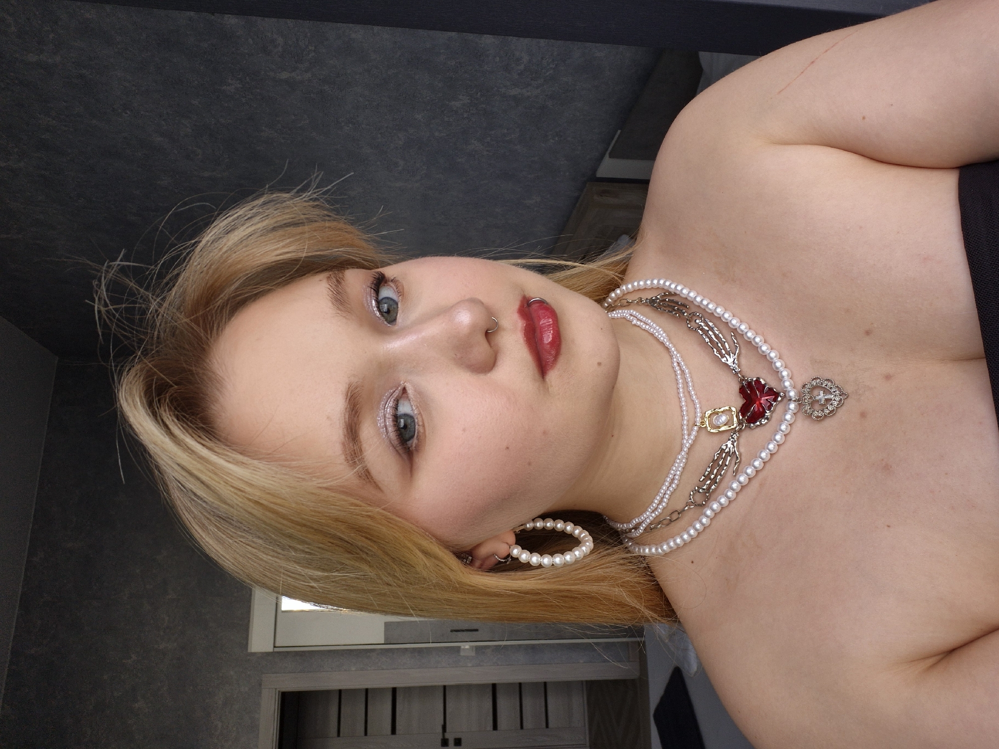 A young Caucasian woman with blonde hair and blue eyes is pictured from the chest up, looking directly at the camera. She is wearing multiple pearl necklaces, large pearl hoop earrings, and has her nose and lower lip pierced. Her makeup includes shimmery eyeshadow and red lipstick. She appears to be in a bedroom with a grey textured wall and a partially visible doorway and closet in the background. The image has a neutral background, with no specific location in Izhevsk, Russia, or any identifying landmarks evident.