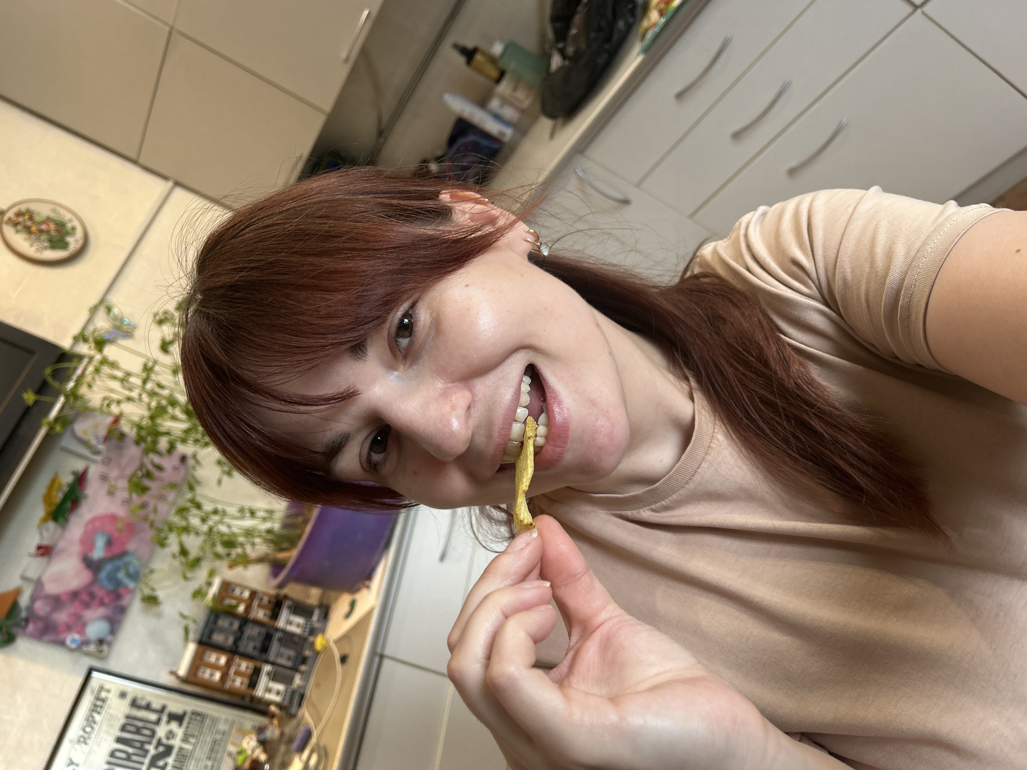 A woman with shoulder-length reddish-brown hair, wearing a beige t-shirt, holds a yellow crispy snack in her hand, positioning it partially in her mouth, seemingly about to take a bite. She is smiling at the camera. Her right hand is holding the snack and has short fingernails with clear nail polish. She has fair skin and a slight mole near her right collarbone. The background shows a kitchen setting with white cabinets and drawers. A plant in a purple pot sits on the counter to her left, along with other items, including a decorative Harry Potter framed poster. The lighting is bright, suggesting an indoor setting. There is no explicit indication of Фоки, Russia.