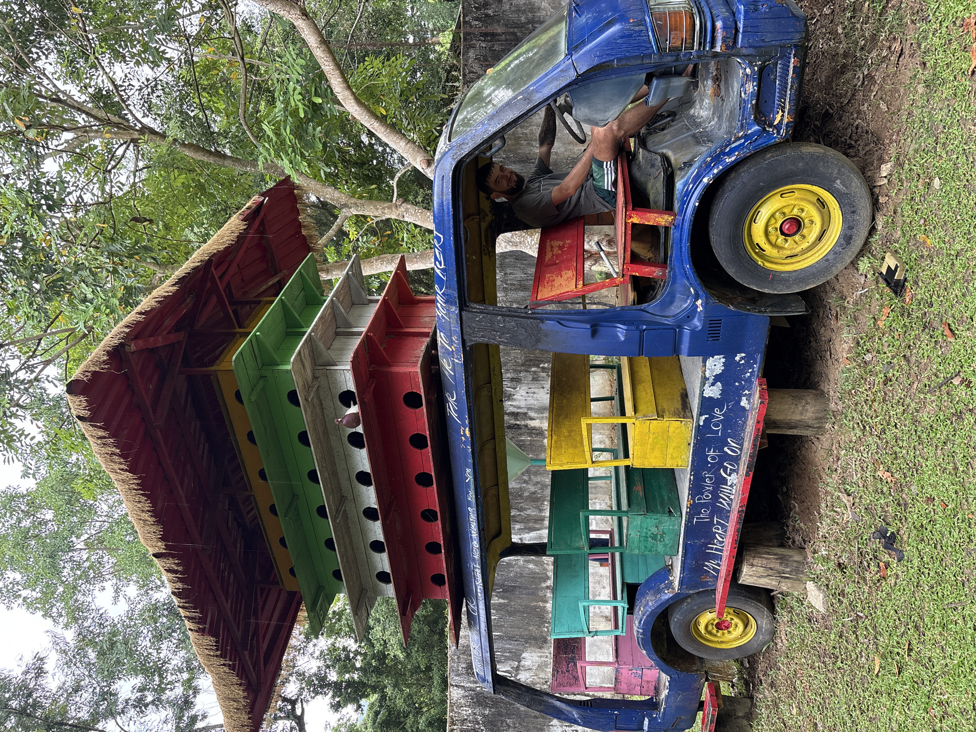 A man sits in the driver's seat of a blue, repurposed truck in Khánh Vĩnh, Vietnam. The truck has been converted into an art installation with colorful tiers extending upward, resembling a multi-level structure with a thatched roof. The tiers are adorned with birdhouse-like structures, each painted a different color. The man in the driver's seat has a beard and appears to be looking at the viewer. The truck is parked on a grassy area. White writing is visible on the side of the truck. The wheels are yellow.