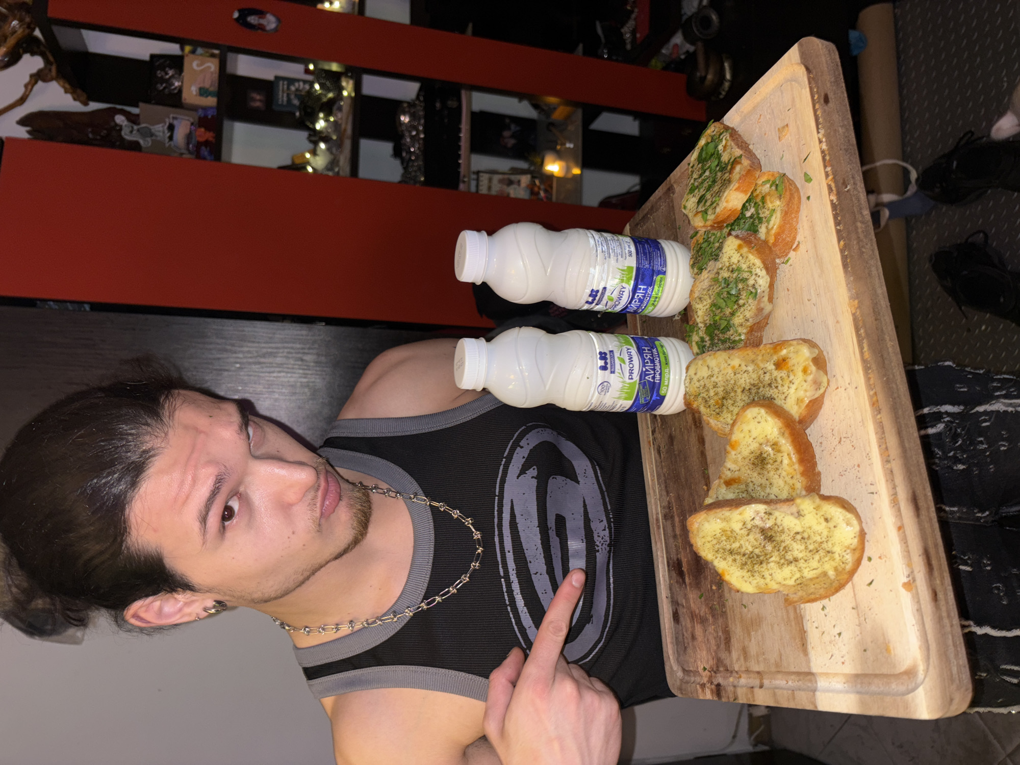 A man with his hair in a bun wearing a black tank top and a silver chain necklace poses with a wooden cutting board holding garlic bread and two bottles of 'AIRYAN' Probiotic dairy product. The background includes a red and black wall, shelving with various items, and a glimpse of patterned clothing and shoes on the floor. The image appears to be taken indoors, but the specific location in Sofia, Bulgaria, cannot be determined from the image alone.