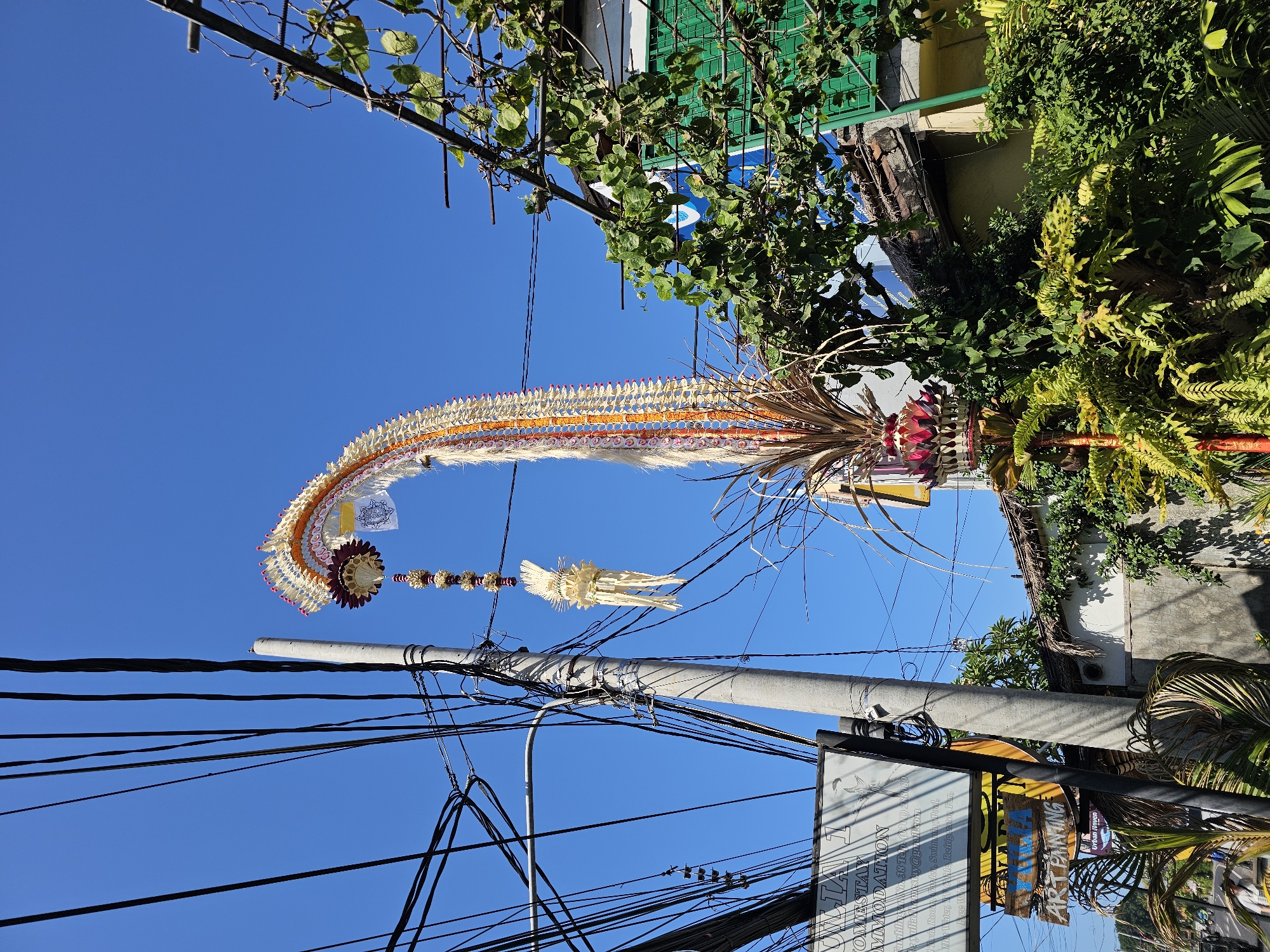 A tall, ornate Balinese Penjor decoration, made of woven leaves and flowers, arches over a street in Denpasar, Indonesia. The Penjor is adorned with intricate patterns and hangs from a utility pole amidst a dense network of electrical wires. In the background, a building with a green-latticed window and lush vegetation are visible. The sky is clear and blue. Signs for "Villa 1 Homestay Accommodation" and "Art Painting" are present on the left side of the image.