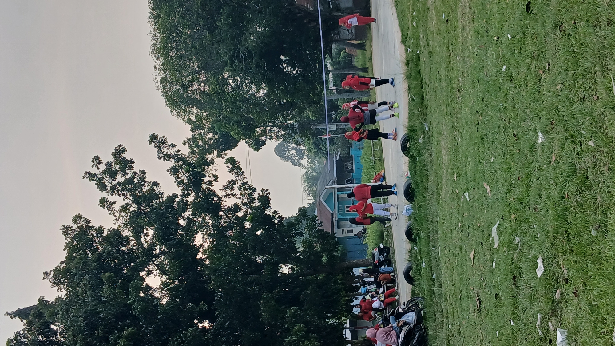 The image depicts an outdoor volleyball court surrounded by grass and trees. Several individuals, predominantly wearing red or dark clothing with some in red headscarves, are present on and around the court. Some are actively playing volleyball, while others are observing or standing by. A volleyball net is visible spanning the court. In the background, a light blue building with a red roof is situated among trees. Several motorcycles are parked near the edge of the court, and there are also some old tires serving as a boundary. The sky is overcast. The scene appears to be a community gathering for a game of volleyball in Klumpang Kebun, Indonesia.