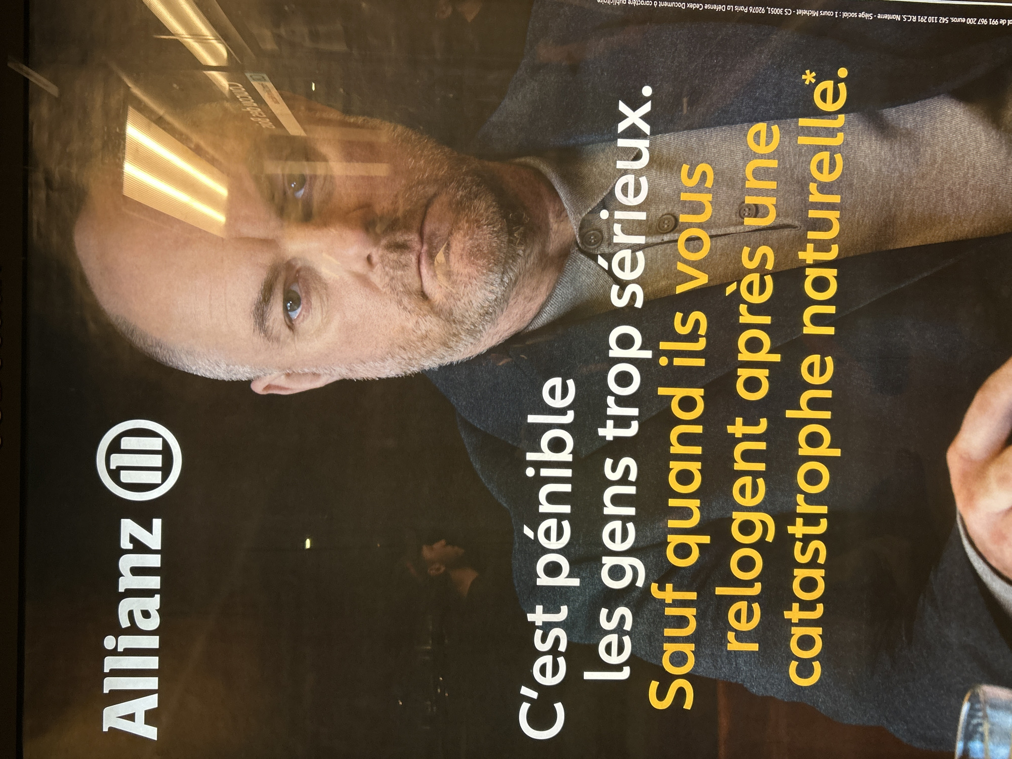 This image is of an Allianz insurance advertisement. The advertisement shows a man with a beard, wearing a button-down shirt and a blazer. The text in the advertisement reads: "C'est pénible les gens trop sérieux. Sauf quand ils vous relogent après une catastrophe naturelle." The Allianz logo is in the bottom left corner. The image also includes some information printed in small text at the top, including references to financial details, the company's location in Paris, France, and that it is a public document. From the reflection, the advertisement appears to be displayed in Strasbourg, France.