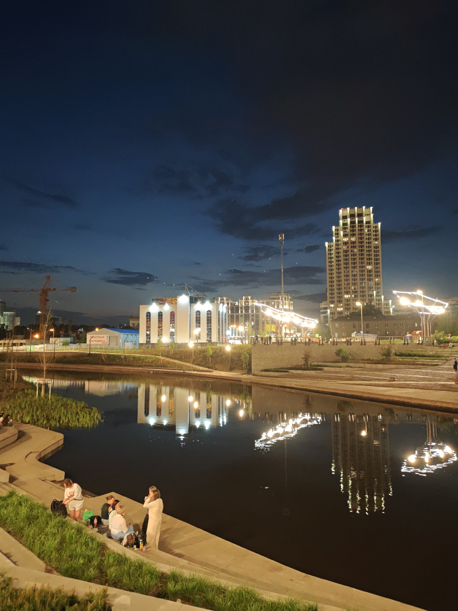 The image shows a night view of Yekaterinburg, Russia. In the foreground, a group of six people is gathered on concrete steps next to a canal. Several bottles are visible near them. Across the canal is an illuminated, white building and a tall, lit skyscraper, both reflected in the water. Streetlights are also reflected in the canal. A construction crane is visible in the background on the left. The sky is dark with some visible clouds.