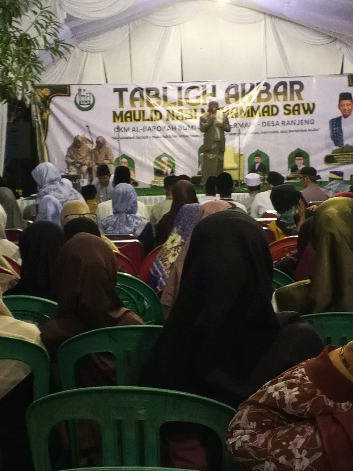 The image captures a religious gathering, likely a "Tabligh Akbar" (Grand Religious Lecture), held under a white tent structure. The backdrop features a large banner with text in Indonesian, indicating the event is in "DKM AL-BAROKAH BUMI..." and mentioning "MAULID NABI MUHAMMAD SAW." On the banner, there are portraits of men, presumably religious figures.
In the foreground, a group of people are seated on green plastic chairs, facing the stage. Many are wearing hijabs or head coverings, typical attire for women in such events in Indonesia. The lighting is somewhat dim, suggesting it might be evening or an indoor event. A person is speaking from a podium on the stage, holding a microphone and a book. Two individuals in traditional religious attire are seated behind the speaker on the stage. The overall atmosphere appears to be one of solemnity and community participation. The banner text suggests the event is related to the celebration of the Prophet Muhammad's birthday and is organized by DKM Al-Barokah in the Bumirangkas/Ranjeng area of Ciruas, Indonesia.