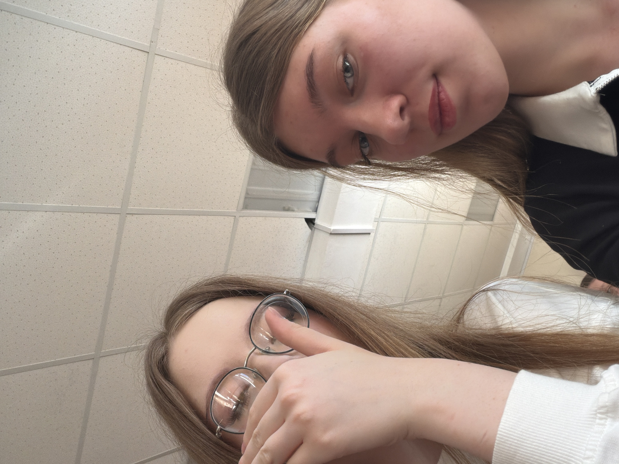 Two young women are posing for a selfie in what appears to be an indoor setting. The first woman has light brown hair, fair skin, and is wearing glasses. She is partially covering her face with her hand. The second woman has light brown hair, fair skin, and is wearing a black top with a white collar. The ceiling is visible in the background and appears to be made of square panels. There is no discernible information in the image to definitively determine the location as Chaikovsky, Russia. The setting appears to be a generic office or institutional building.
