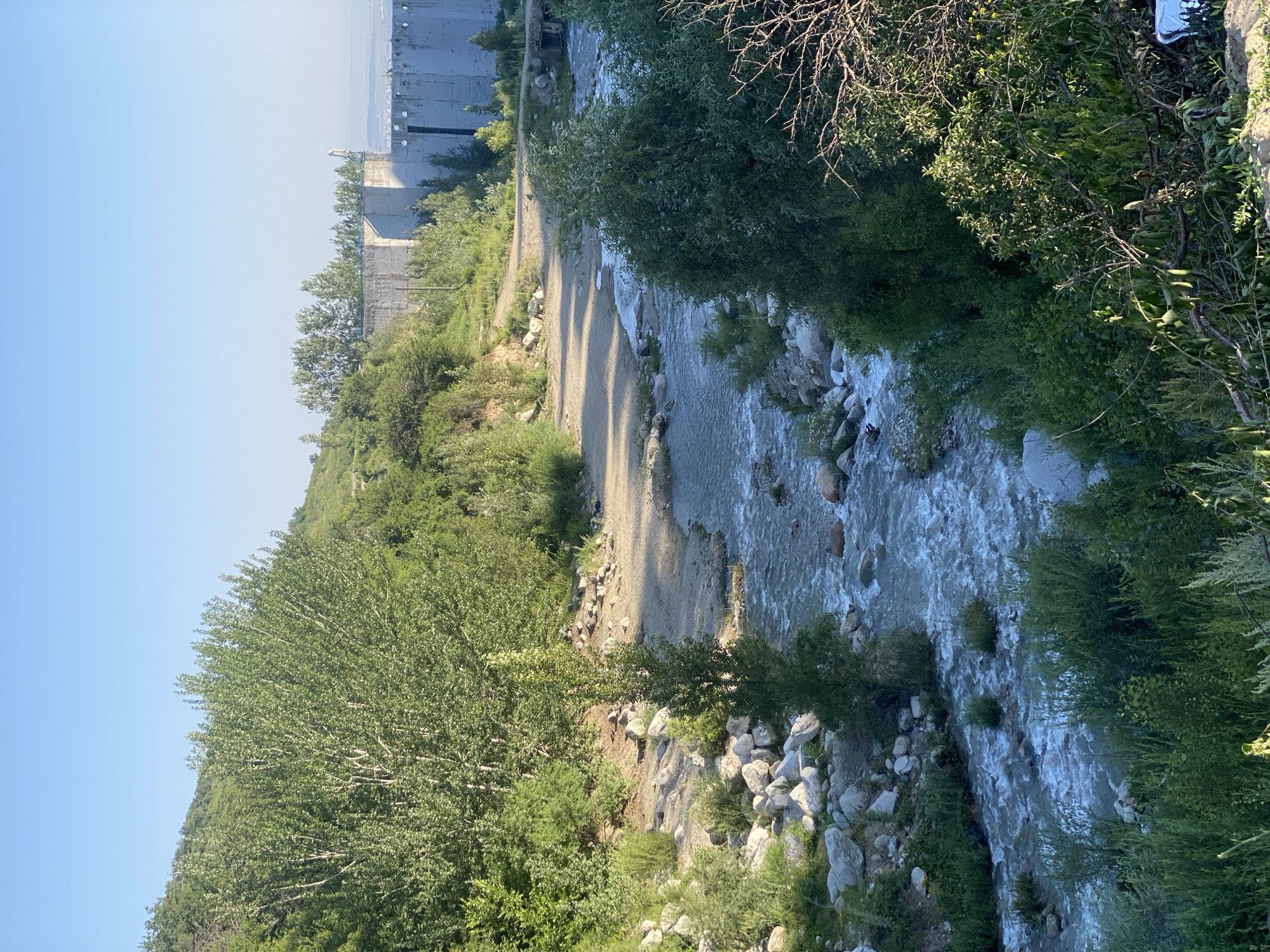 The image captures a scenic view of a rocky river flowing through a verdant, hilly landscape under a clear blue sky. On the right side of the image, a large concrete structure, likely a dam or hydroelectric power station, dominates the background. The dam is a series of massive, light-colored concrete blocks, with some sections appearing weathered. Trees and lush green vegetation surround the river and climb the hills. The river itself is a central feature, with clear, flowing water over a bed of smooth stones and rocks. A sandy path runs alongside the river, leading towards the concrete structure. The overall impression is one of a natural environment integrated with man-made infrastructure. This scene is located in the foothills surrounding Almaty, Kazakhstan, specifically near the **Medeu Dam**, which is part of the complex that includes the Medeu high-mountain skating rink and the Shymbulak ski resort.