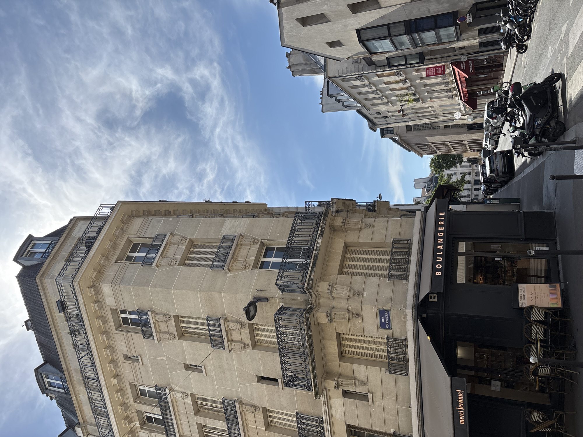 The image captures a street scene in Paris, France, likely in the Latin Quarter, given the presence of the "Rue Mouffetard" street sign. The left side of the frame is dominated by a corner building with a light-colored stone facade, featuring multiple stories, ornate balconies with black metal railings, and numerous windows with closed wooden shutters. A black awning extends from the ground floor, advertising "merci jerome!" and "BOULANGERIE". Outside the boulangerie, tables and chairs are set up for outdoor seating.
The street itself is narrow and slopes slightly downhill. Parked cars and scooters line both sides of the street. The buildings on the right side of the street are also multi-story, with varied architectural styles, including a building with a modern glass facade and another with a red awning. The sky is bright blue with wispy white clouds. The overall impression is a typical Parisian street on a sunny day, with a focus on urban architecture and daily life.