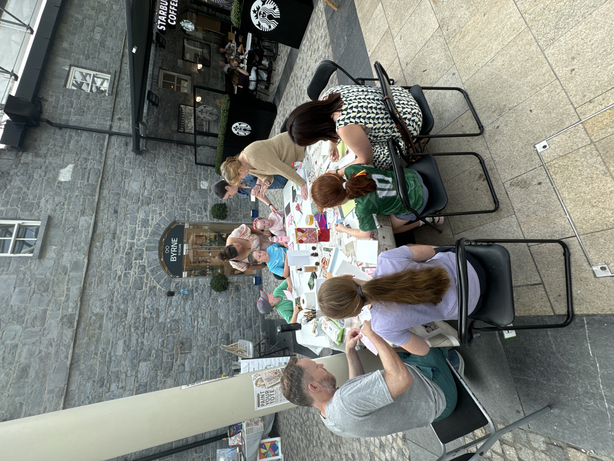 A group of people are gathered around a table outdoors, engaged in a craft activity. The setting appears to be a public space in Kilkenny, Ireland, likely near a Starbucks as indicated by the visible signage. The table is covered with various craft supplies, including paper, scissors, glue, paint, and brushes. Several individuals, including adults and children, are actively participating, with some cutting paper and others painting. A man in a grey t-shirt is focused on his work, as is a young girl with red hair wearing a green and white soccer jersey. The background reveals stone buildings and a covered area, suggesting a sheltered outdoor space or courtyard. The overall atmosphere is one of creative engagement and community.