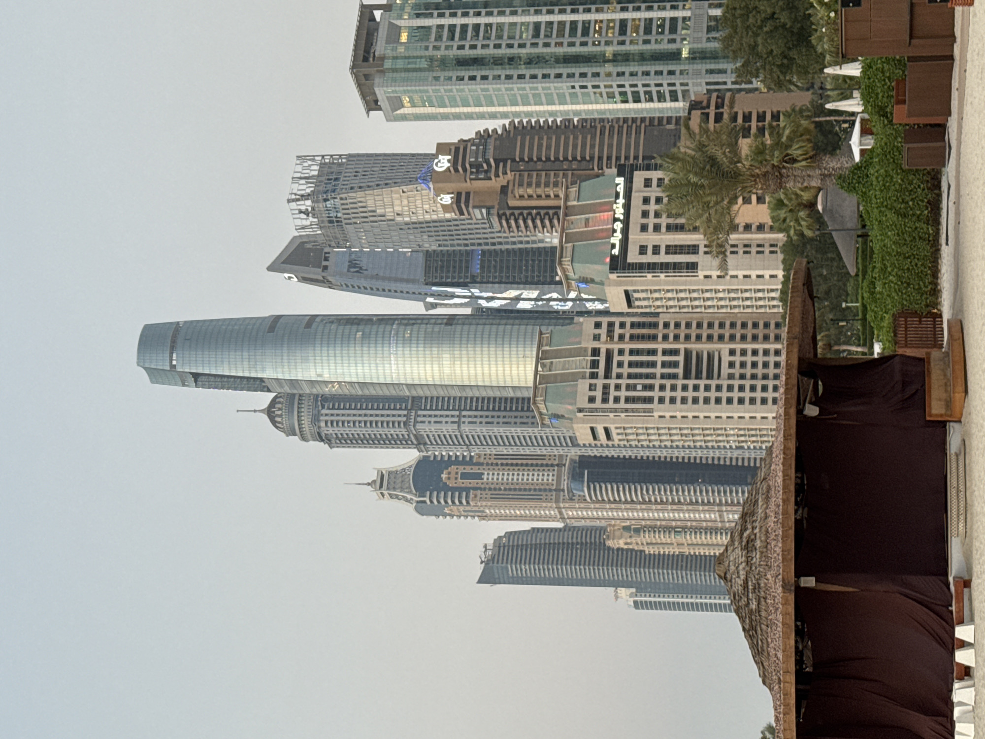 The image captures a panoramic view of the Dubai Marina skyline, a vibrant district in Dubai, United Arab Emirates. The skyscrapers are a prominent feature, with a particularly tall, slender, and curved glass skyscraper dominating the center of the frame. Several other high-rise buildings, characterized by their modern architecture and varying shapes and colors, flank this central structure. Some buildings exhibit intricate facade designs with numerous windows, while others have a sleeker, more contemporary appearance. In the foreground, a covered structure with a thatched roof and dark brown fabric walls sits on a sandy surface, likely a beach cabana. The sandy ground extends towards the viewer, with hints of white beach chairs and some wooden structures visible. Lush green bushes and trees, including a date palm, are scattered across the middle ground, adding a touch of nature to the urban landscape. The sky above is overcast and pale, contributing to a soft, diffused light. No people are visible in the scene.