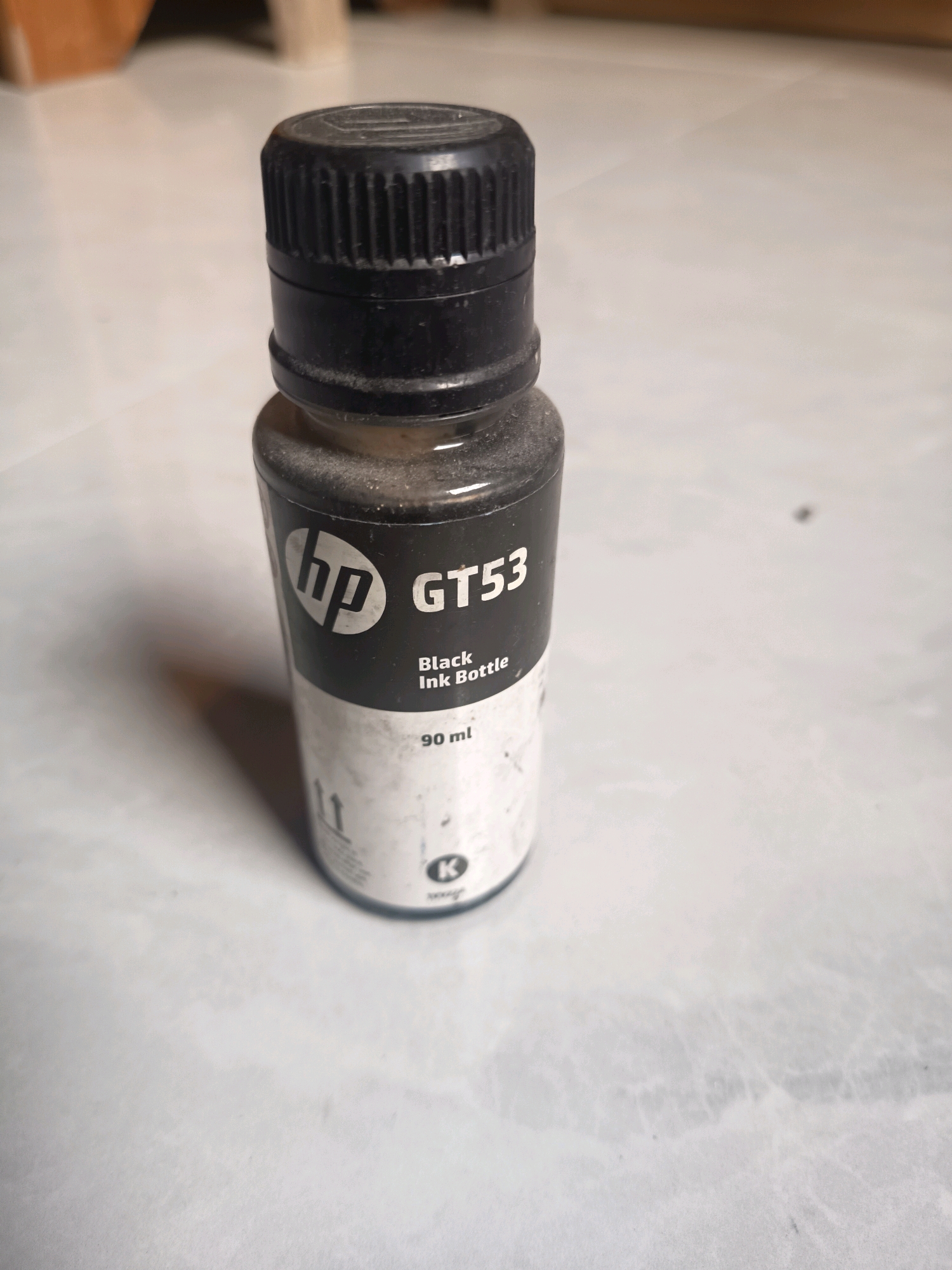 The image shows an HP GT53 black ink bottle with a black cap, sitting on a tiled surface. The bottle is partially filled with black ink and has labels indicating it is "Black Ink Bottle" and "90 ml". There is a "K" symbol in a circle on the lower part of the label. The background appears to be a blurred indoor environment with some wooden furniture. The image does not provide any information about the city of Sidorejo, Indonesia, or any specific location within it. There are no people or actions depicted in the image.