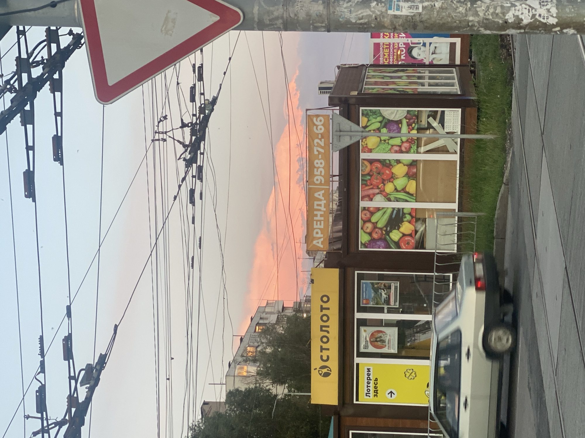 The image depicts a street scene in Samara, Russia, during sunset. The sky is filled with pink and orange clouds. In the foreground, a light-colored sedan is captured in motion, driving on a street with tram tracks. To the right of the car is a kiosk with a yellow awning displaying the "СТОЛОТО" logo. Next to it, another kiosk has an orange awning with the word "АРЕНДА" and a phone number. The kiosk windows are decorated with images of fruits and vegetables. Overhead, a complex network of trolleybus wires crisscrosses the frame. A large, inverted red and white triangular yield sign is prominently visible on the right side of the image. Further back, an apartment building with illuminated windows can be seen. The overall atmosphere is urban, with a touch of natural beauty from the sunset.