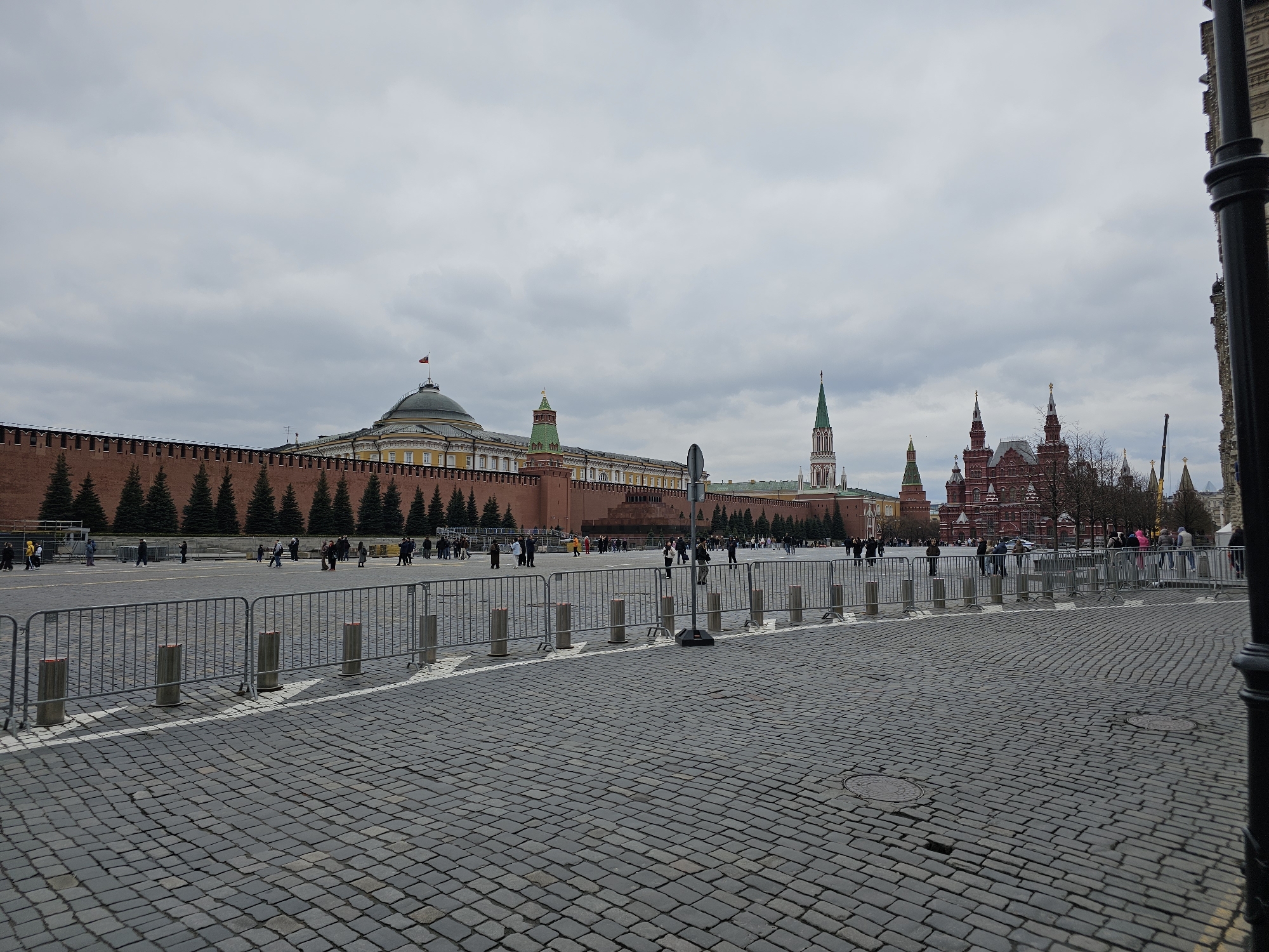 The image depicts a view of Red Square in Moscow, Russia on a cloudy day. In the foreground is a cobblestone pavement, partially obscured by a metal barricade. Beyond the barricade, there are several people walking around in the open area. In the background, a long red brick wall with evergreen trees planted along it is visible. Further back, there are several buildings, including the Kremlin with its various towers and domes. The overall impression is a busy public space in a historic area of the city.