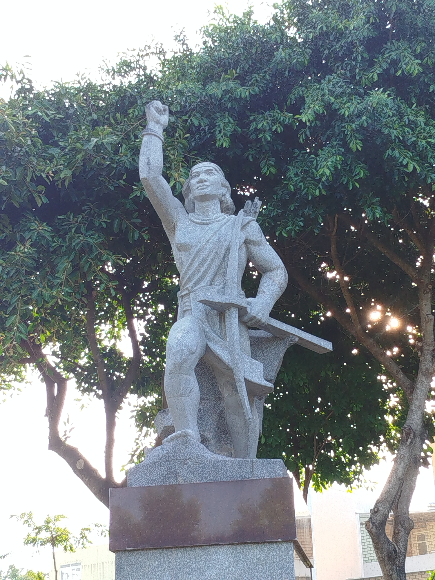 A full shot captures a gray stone statue, possibly depicting a historical figure, positioned on a platform in Pleiku Ward, Vietnam. The figure is male, muscular, and dressed in a tunic or robe. His right arm is raised in a fist, while his left arm holds a long, flat object. A bow and quiver are visible on his back. The statue is set against a backdrop of a large tree with dense foliage, with sunlight filtering through the leaves. In the background, parts of a building are visible.