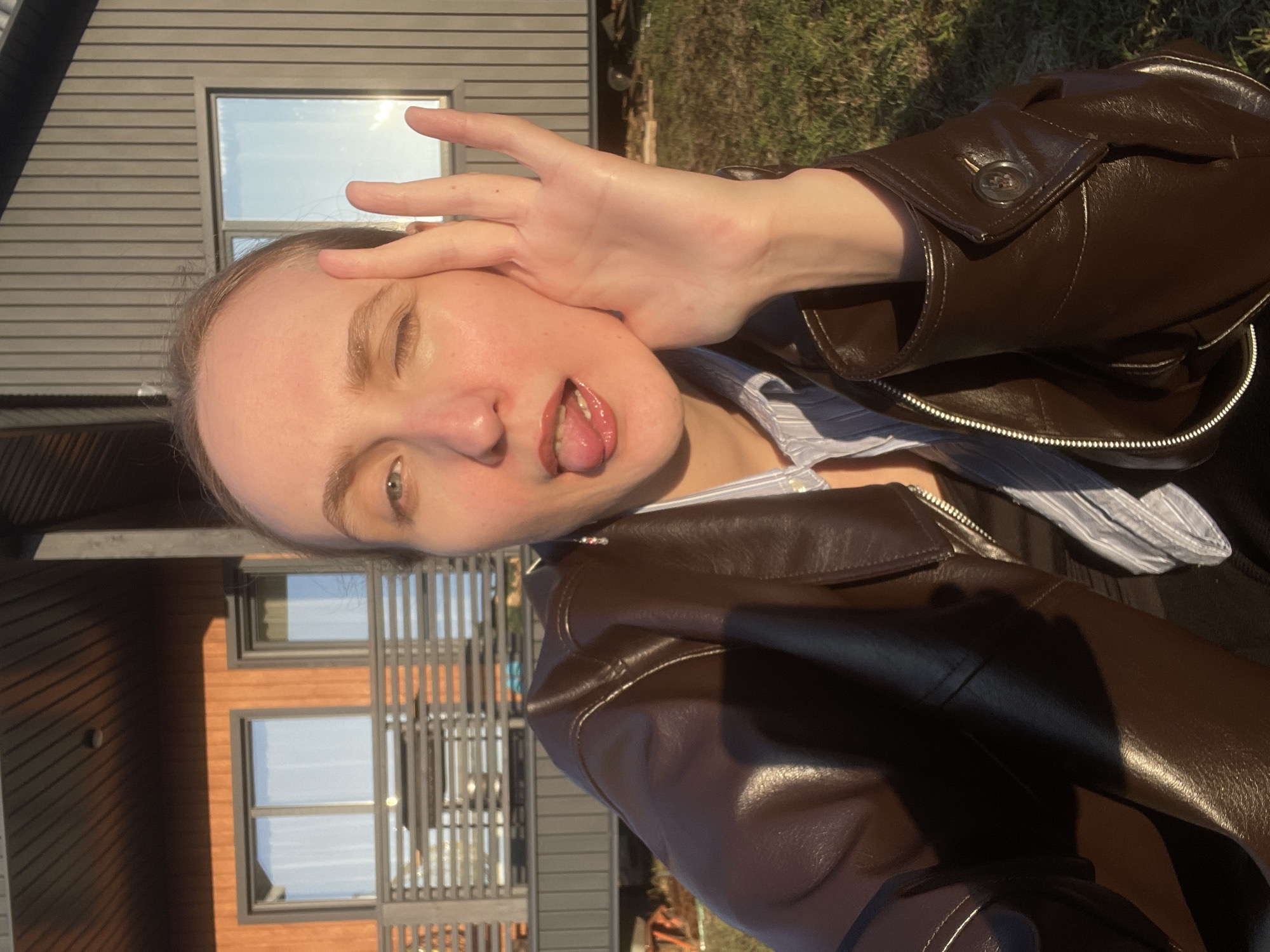 An outdoor, rotated close-up portrait captures a young person making a playful face on a sunny day. The subject, a light-skinned individual with light hair, is positioned in the foreground, looking directly at the viewer with their head slightly tilted. They have their tongue partially extended and one eye squinted in a humorous expression. Their right hand is raised with fingers splayed, covering part of their forehead and temple, possibly shielding from the bright sunlight. They are wearing a dark brown, possibly leather or faux leather, zippered jacket over a light-colored, subtly striped collared shirt.
The background reveals a modern building with gray horizontal siding and dark-framed windows. Reflections in the windows indicate a clear, bright sky. To the right of the subject, green grass is visible, confirming an outdoor setting. The strong, direct sunlight suggests the time of day is either morning or late afternoon. The overall mood is candid and lighthearted. There is no visible text in the image.