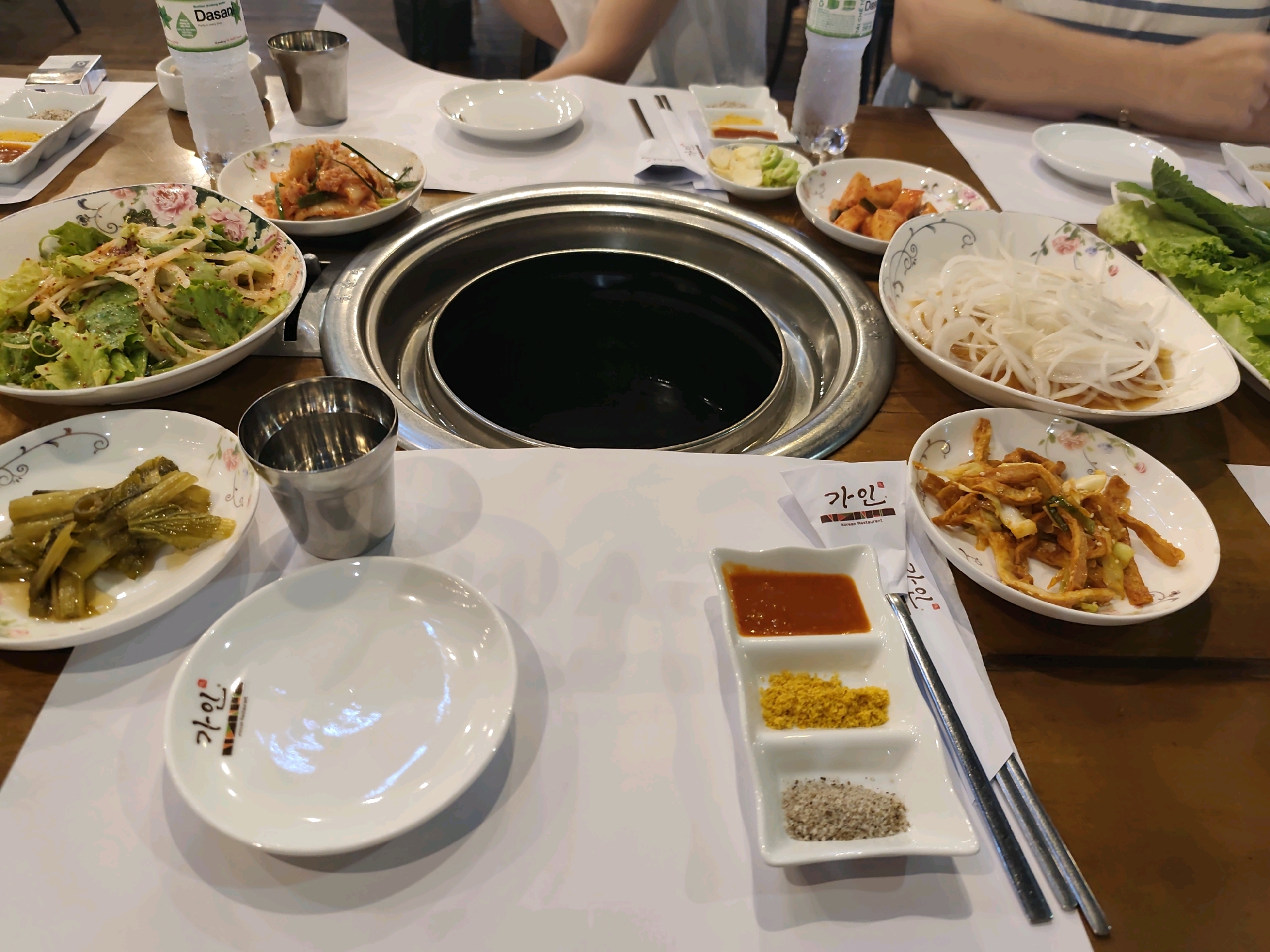 The image depicts a table set for a meal, likely Korean barbecue, in an indoor setting. A circular grill is centrally placed on the wooden table, surrounded by various dishes of food. To the left, a large bowl contains a salad with lettuce and thinly sliced onions, dressed with a light sauce. Next to it, a smaller oval dish holds kimchi. In front of the grill are three small white condiment dishes, one with a red sauce, another with a yellow powder, and the third with a mixture of salt and pepper. A plate with what appears to be stir-fried food, possibly fish cakes or meat, is positioned to the right of the condiments. Further right, a platter holds lettuce leaves and other green vegetables, likely for wrapping food. A bottle of Dasani water is visible in the background. Several metal cups and a white plate with Korean writing, likely a restaurant logo, are also present. The hands and arms of at least one person are visible in the upper portion of the image, suggesting diners are present. The lighting is natural and appears to be coming from above. The overall impression is a spread of food ready to be consumed.