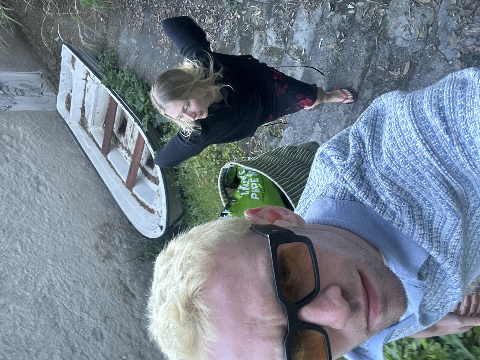 A high-angle, selfie-style shot features two individuals near what appears to be a small, weathered rowboat. The first person, in the foreground, is a light-skinned male with bleached blonde hair and sunglasses, wearing a light blue woven shirt. The second person is a light-skinned female with blonde hair wearing a black top and floral skirt, facing away from the camera. The boat is situated on a stony surface with some vegetation. A striped tote bag with a visible logo in green is partially visible between the two people. Based on available context, the location is consistent with Lipari, Italy.